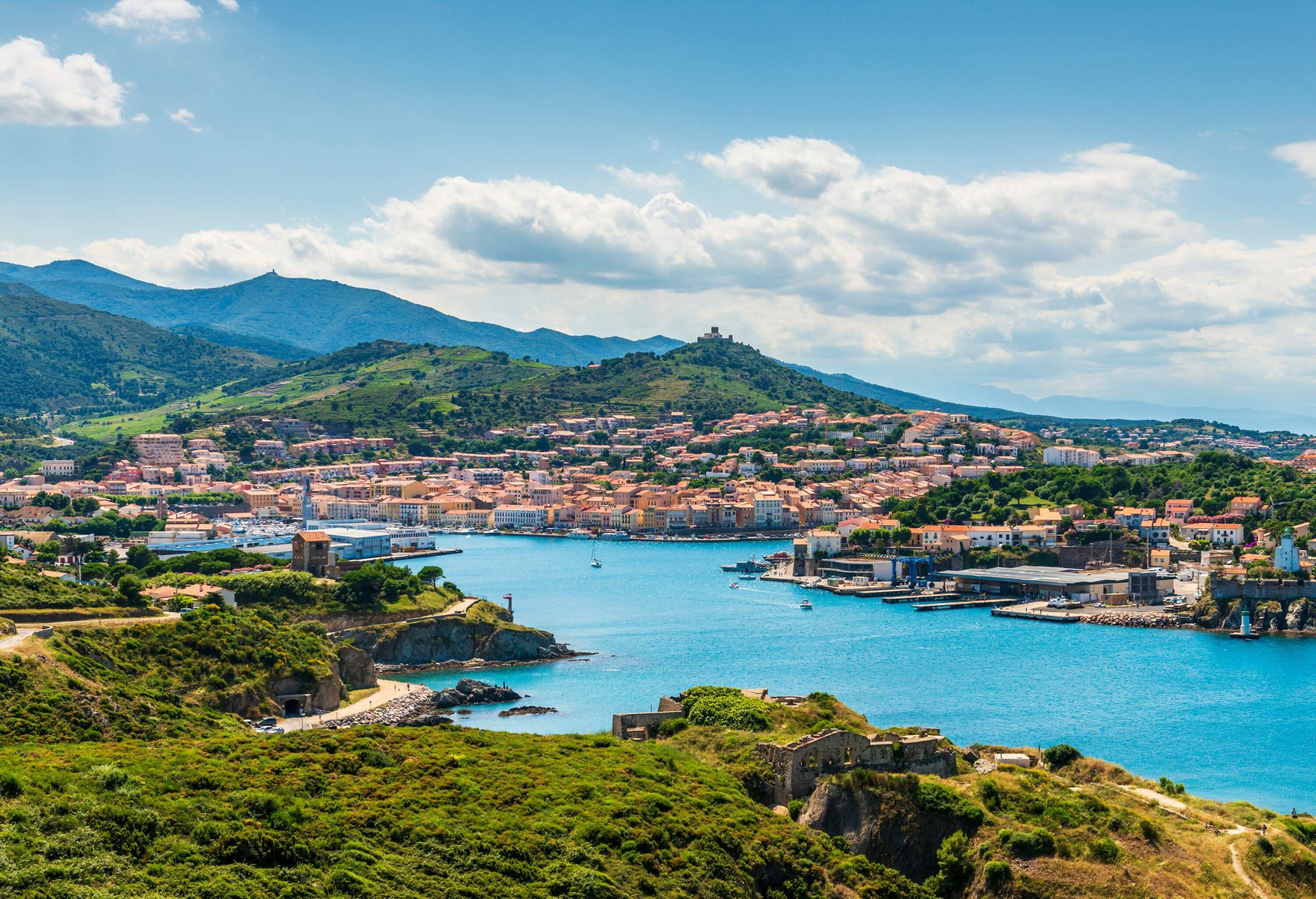 A turquoise sea and a coastal cityscape beneath the lush mountain range against the cloudy blue sky.