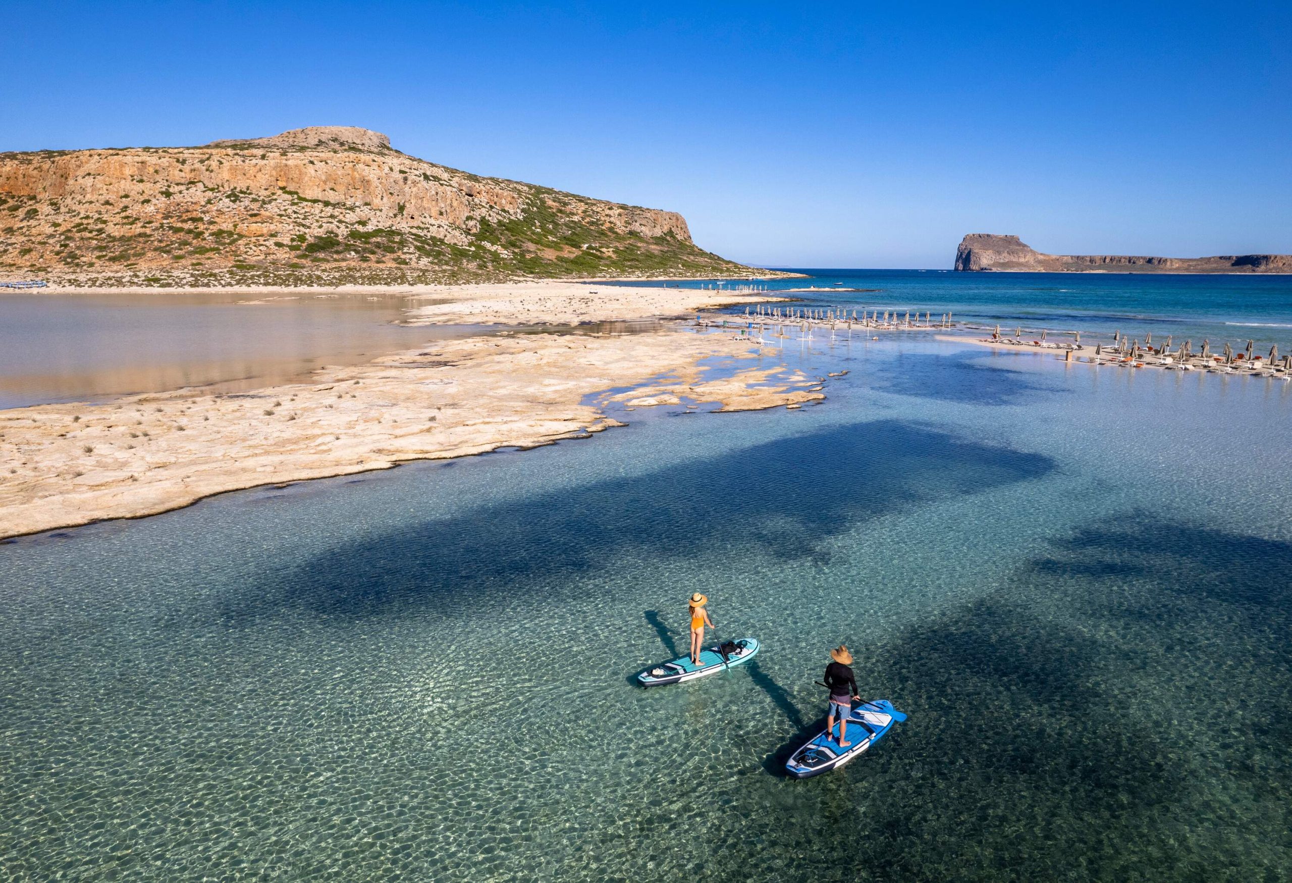A couple stand-up paddle boarding on shallow calm crystalline waters surrounded by rugged cliffs.