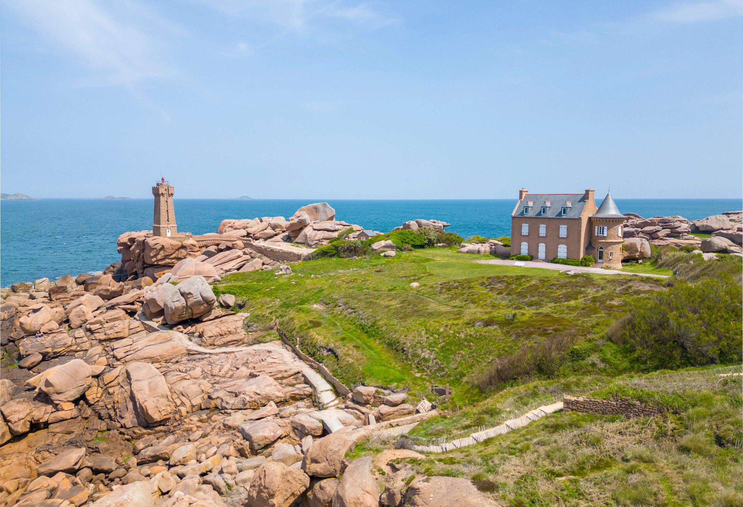 Wide angle view of a hill made or rocks and green grass, with a lighthouse by the sea and a large building