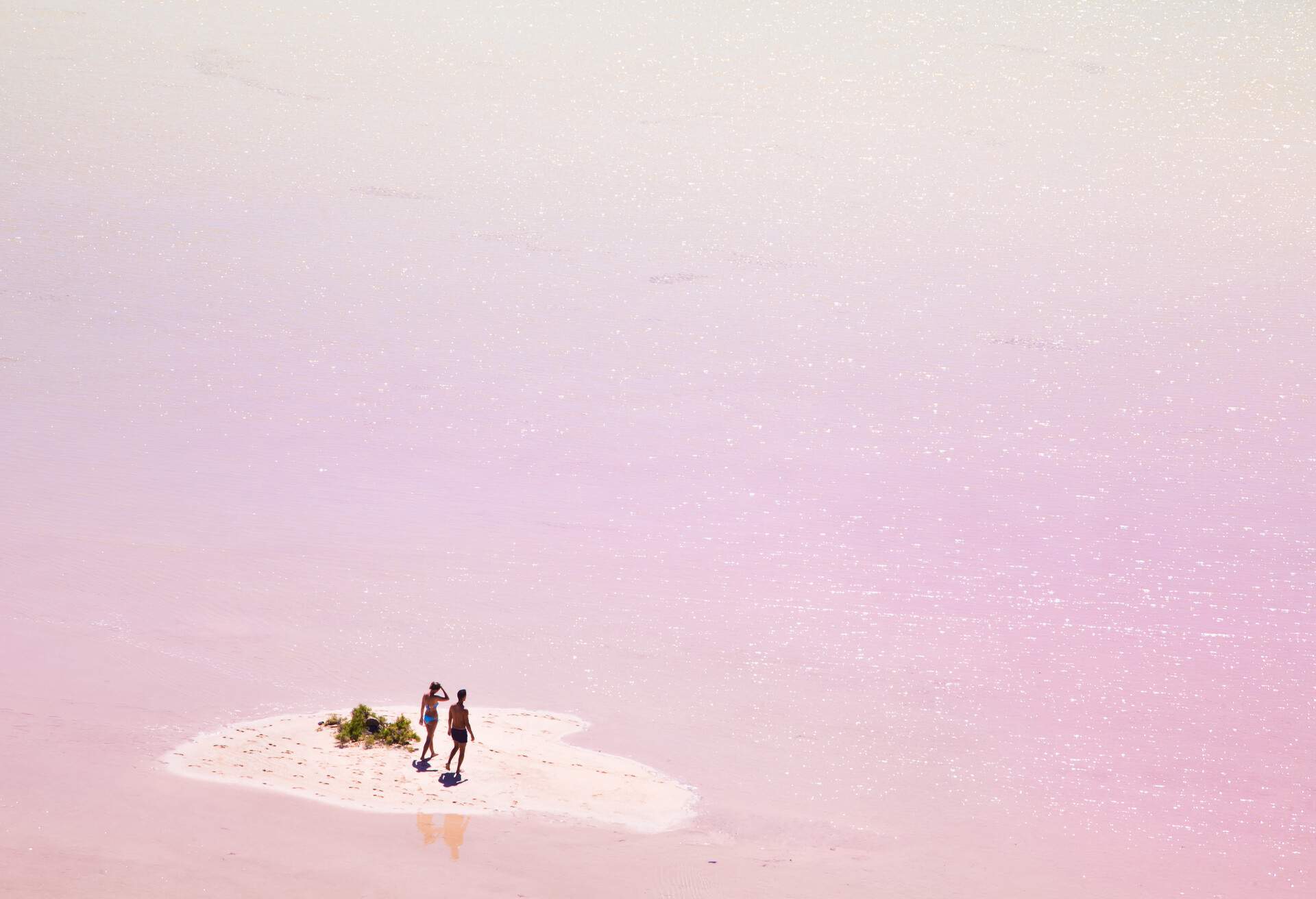 Two people stand on a sandbar of a pink lagoon.