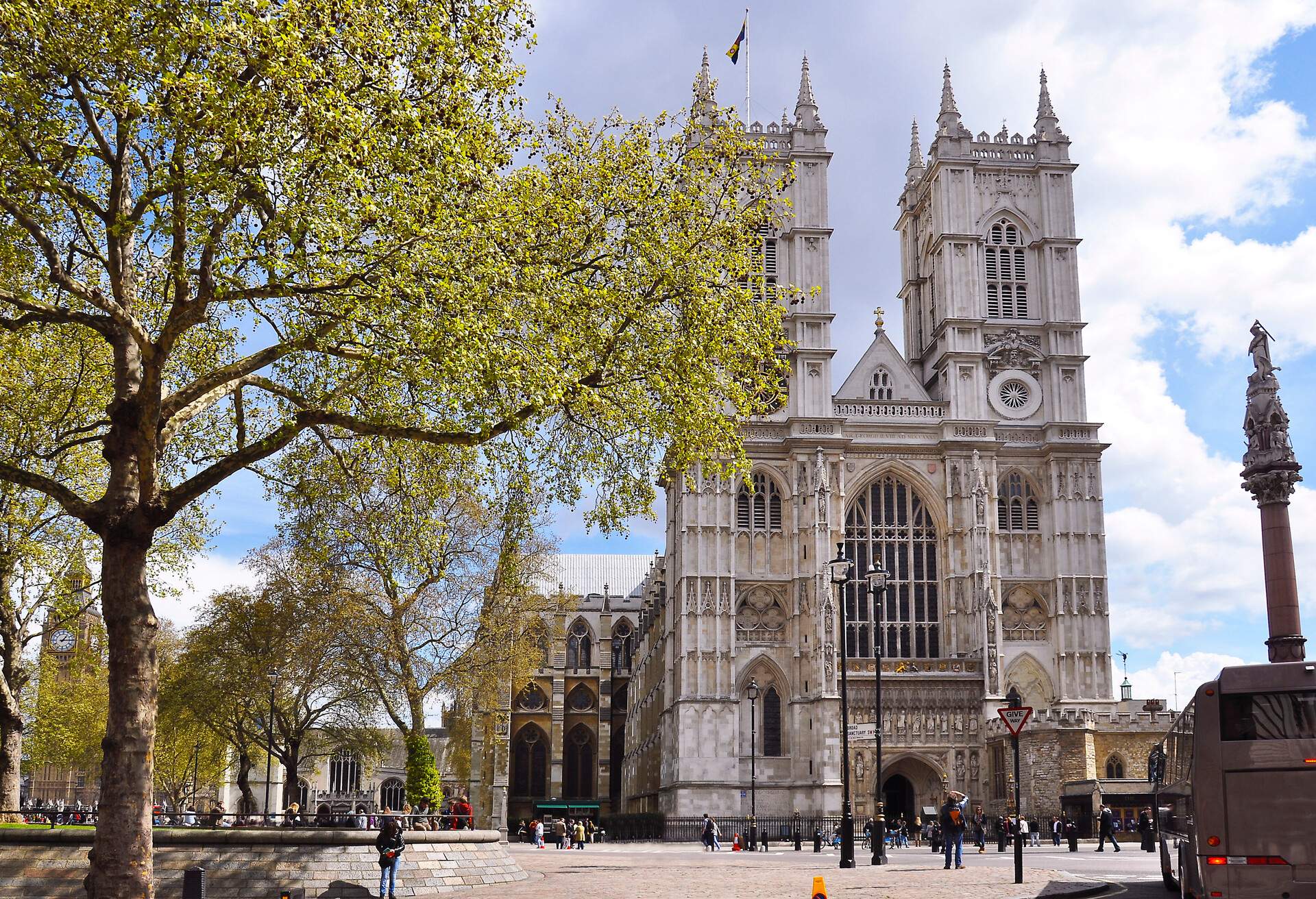 Westminster Abbey is a gothic church with an enormous nave window and twin towers with pinnacles in front of a crowded open space with autumnal trees.