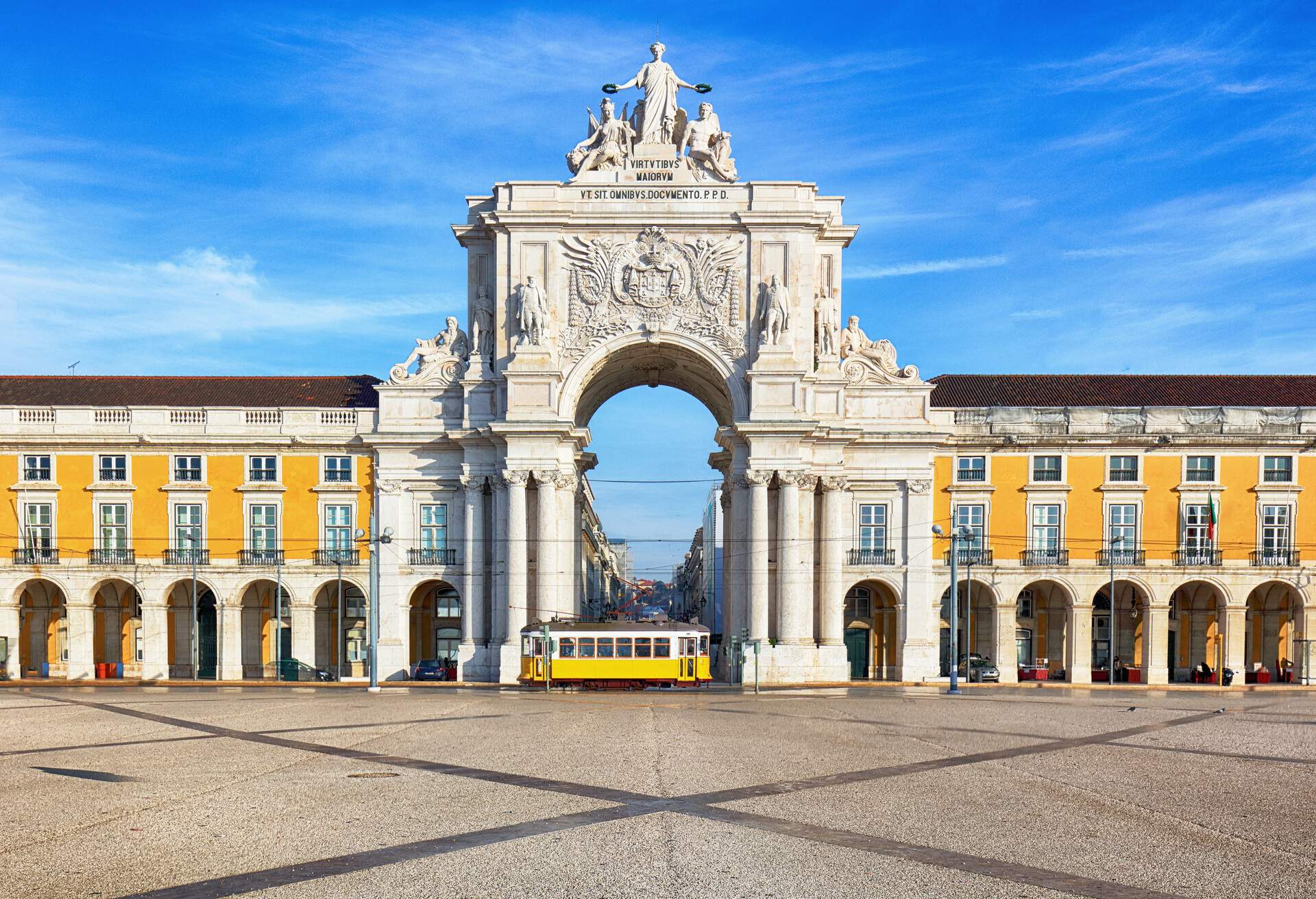 A yellow tram passed the stone memorial arch connecting the arcades supported by columns at regular intervals in a town square.