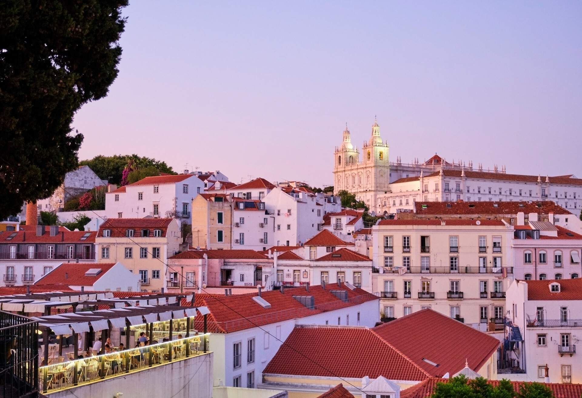 An old city skyline dominated by buildings with orangy tiled roofs and a monastery in the middle against the dramatic sunset sky.