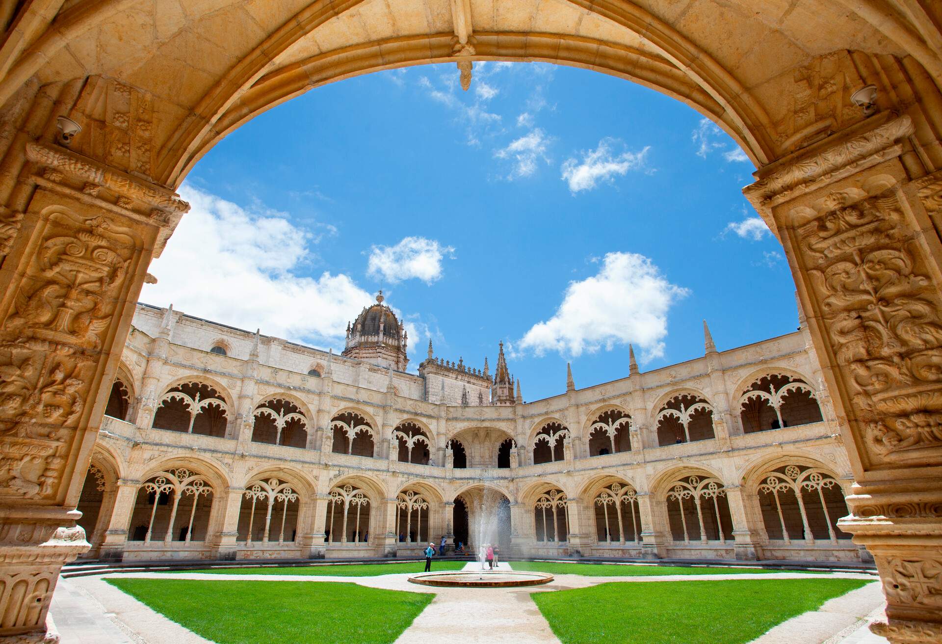 A fountain in the centre of a grass courtyard guarded by ornate cloisters with arch windows.