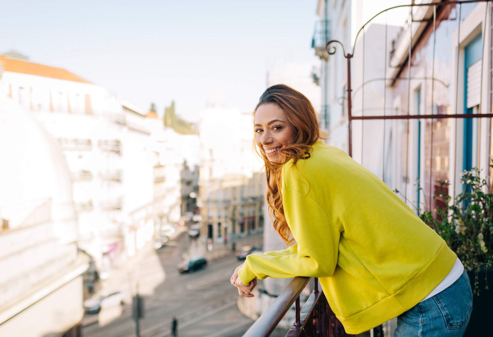 A smiling woman in a mustard jacket leans on a balcony.