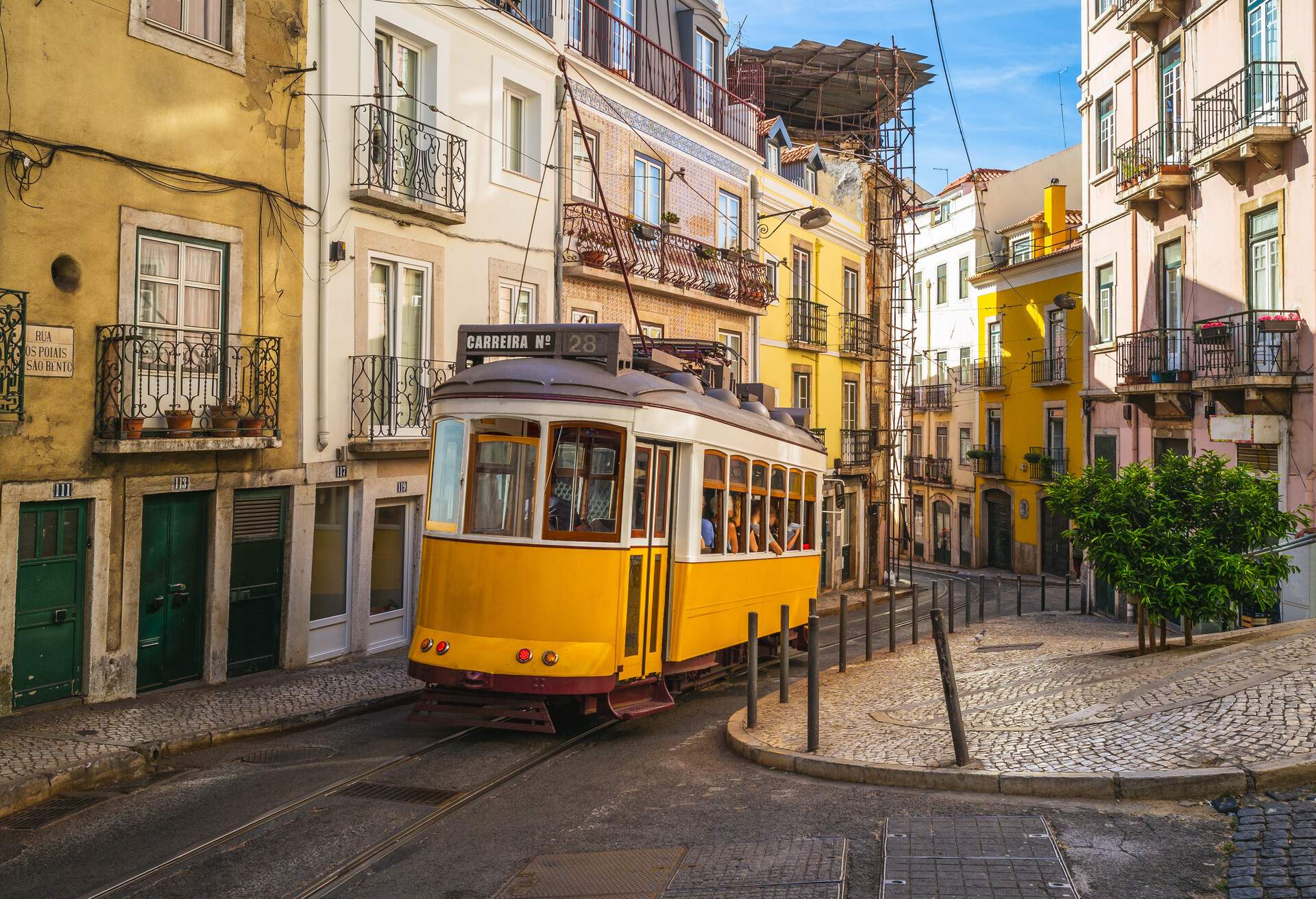 A yellow tram travelling on a narrow railway road flanked by colourful buildings.