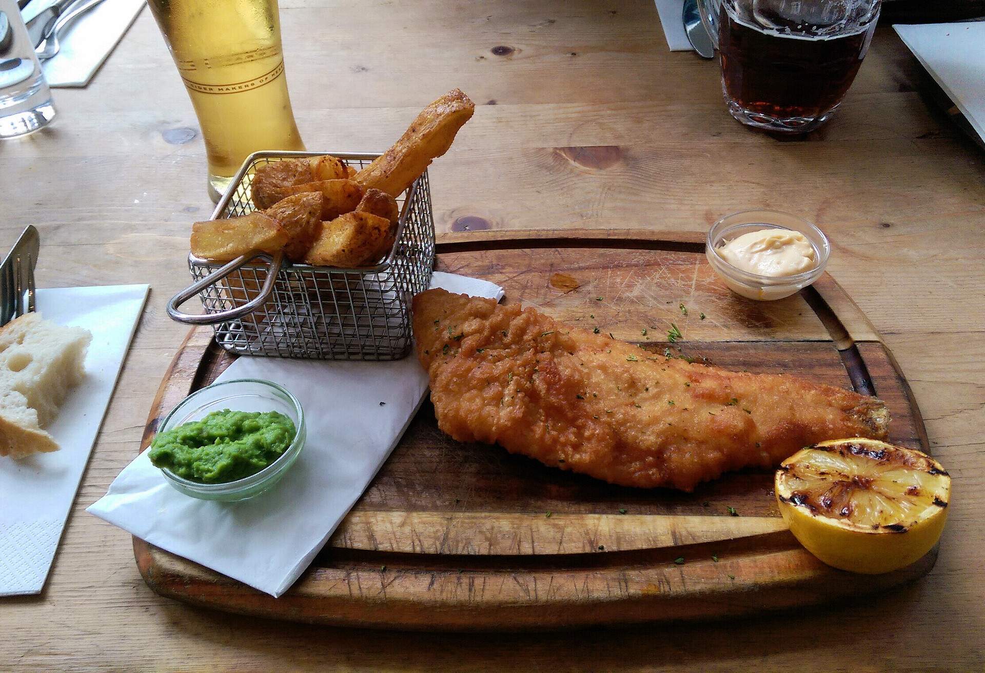 Fish and chips are presented on a wooden board with cutlery and a slice of bread on the side.