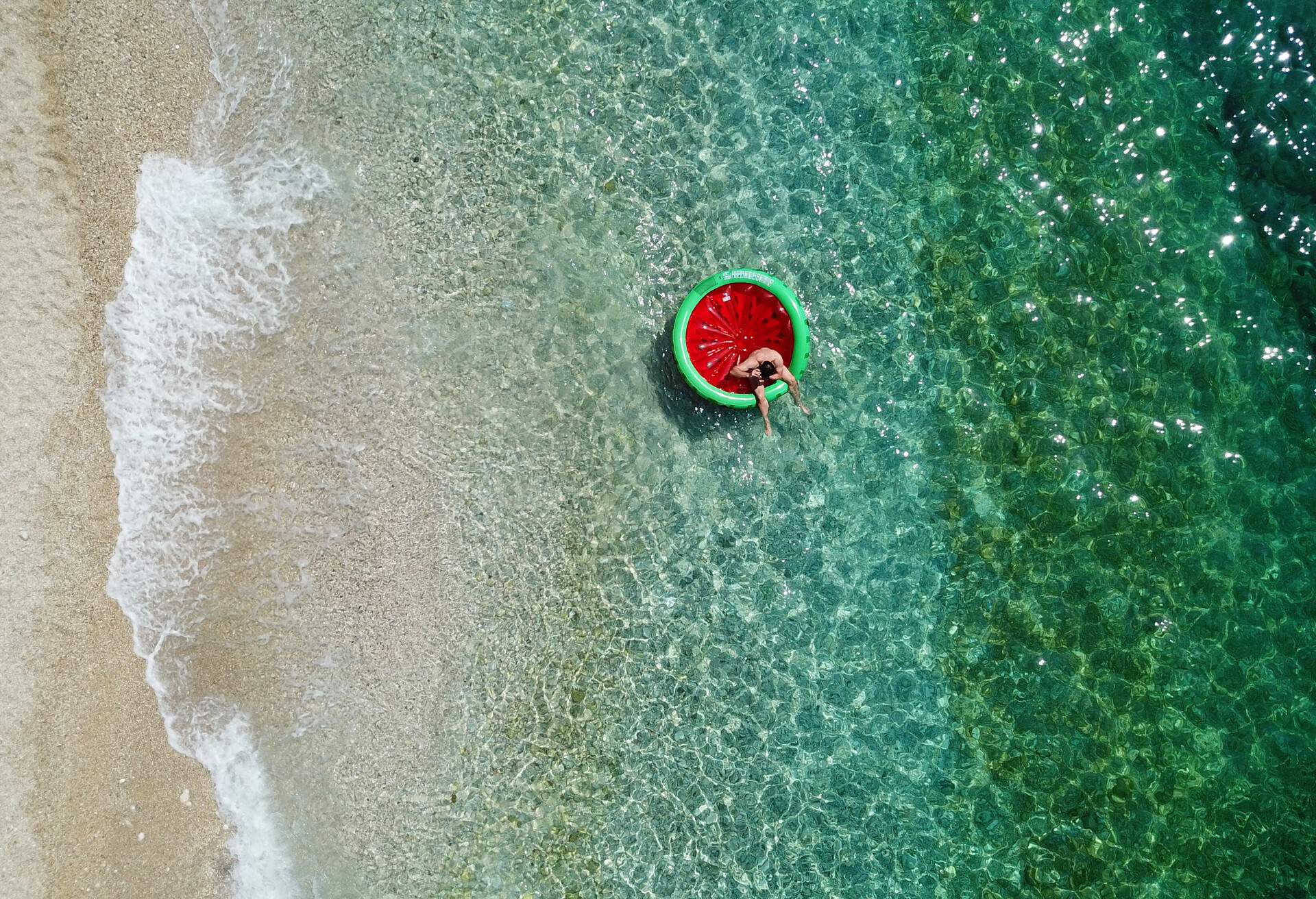 Top view of an individual sitting in an inflatable watermelon on the shore of the emerald sea.