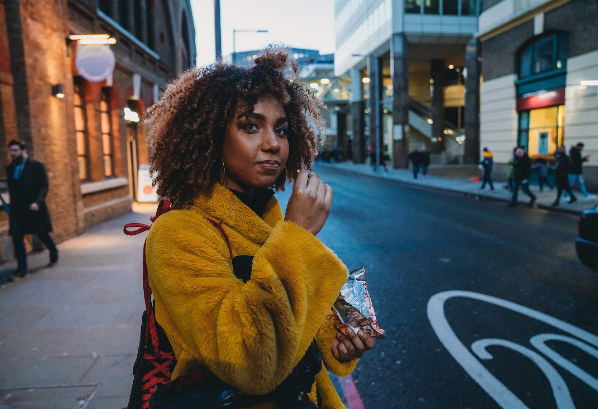 A young woman in a yellow fuzzy coat having a snack while standing on the sidewalk.