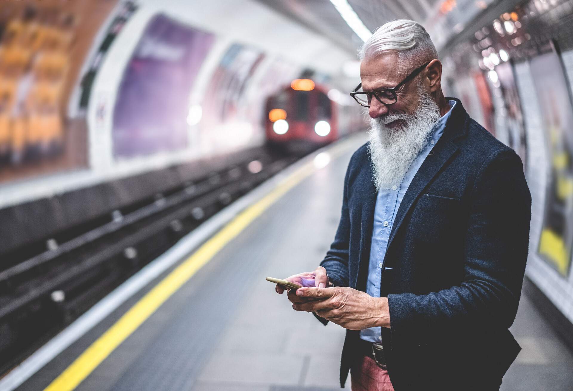 A senior citizen wearing formal attire smiles while glancing at his smartphone in the subway while he waits for the train.