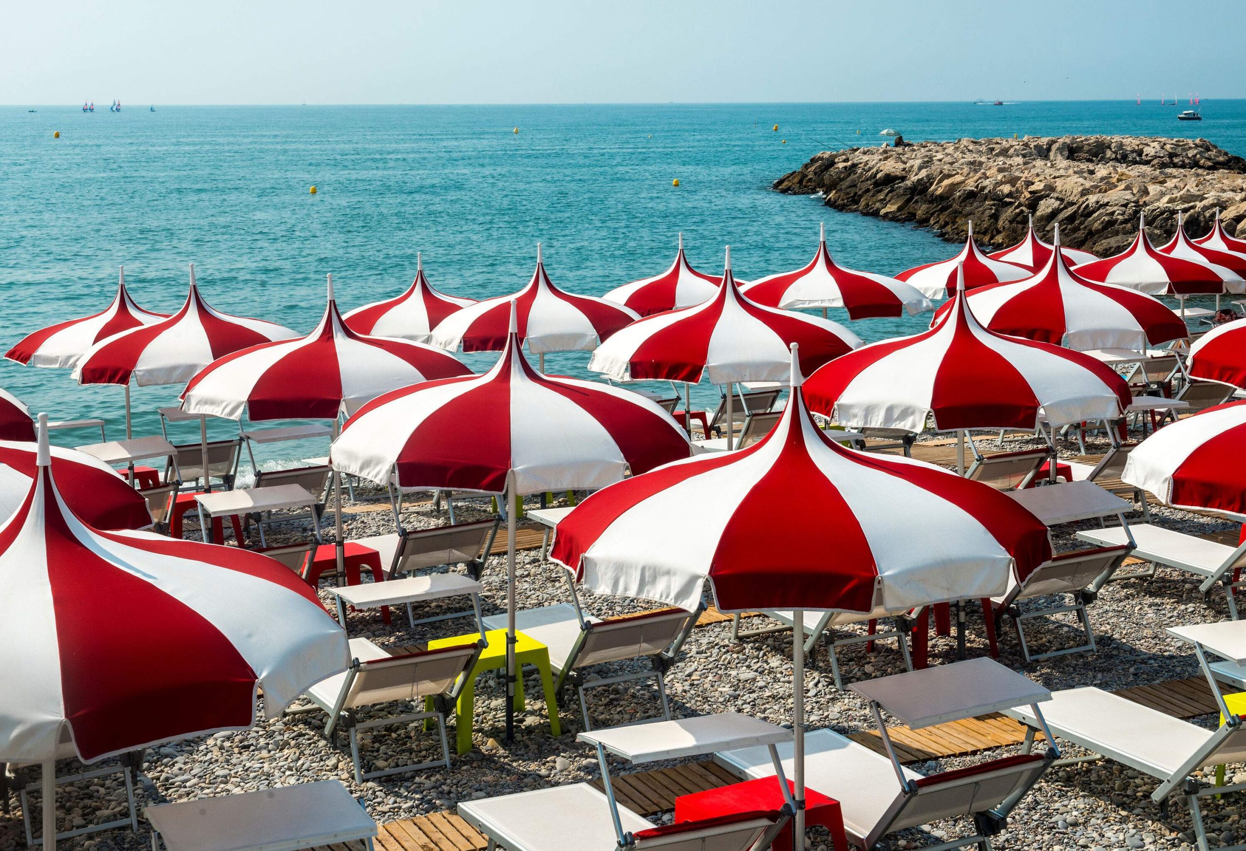Sun loungers with red and white parasols lined up along a stony beach with a panoramic view of the ocean.