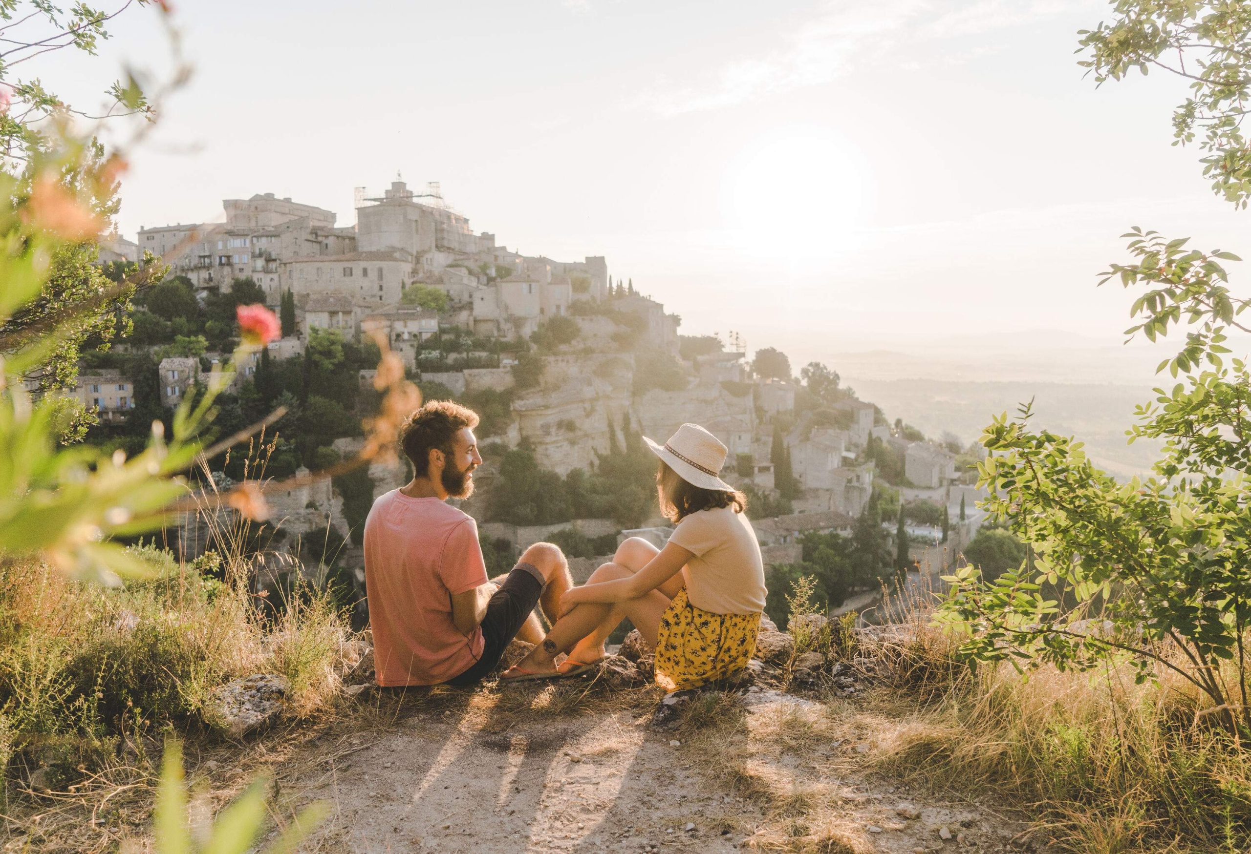 A man and a woman sitting on a sandy cliff across from a hilltop village.