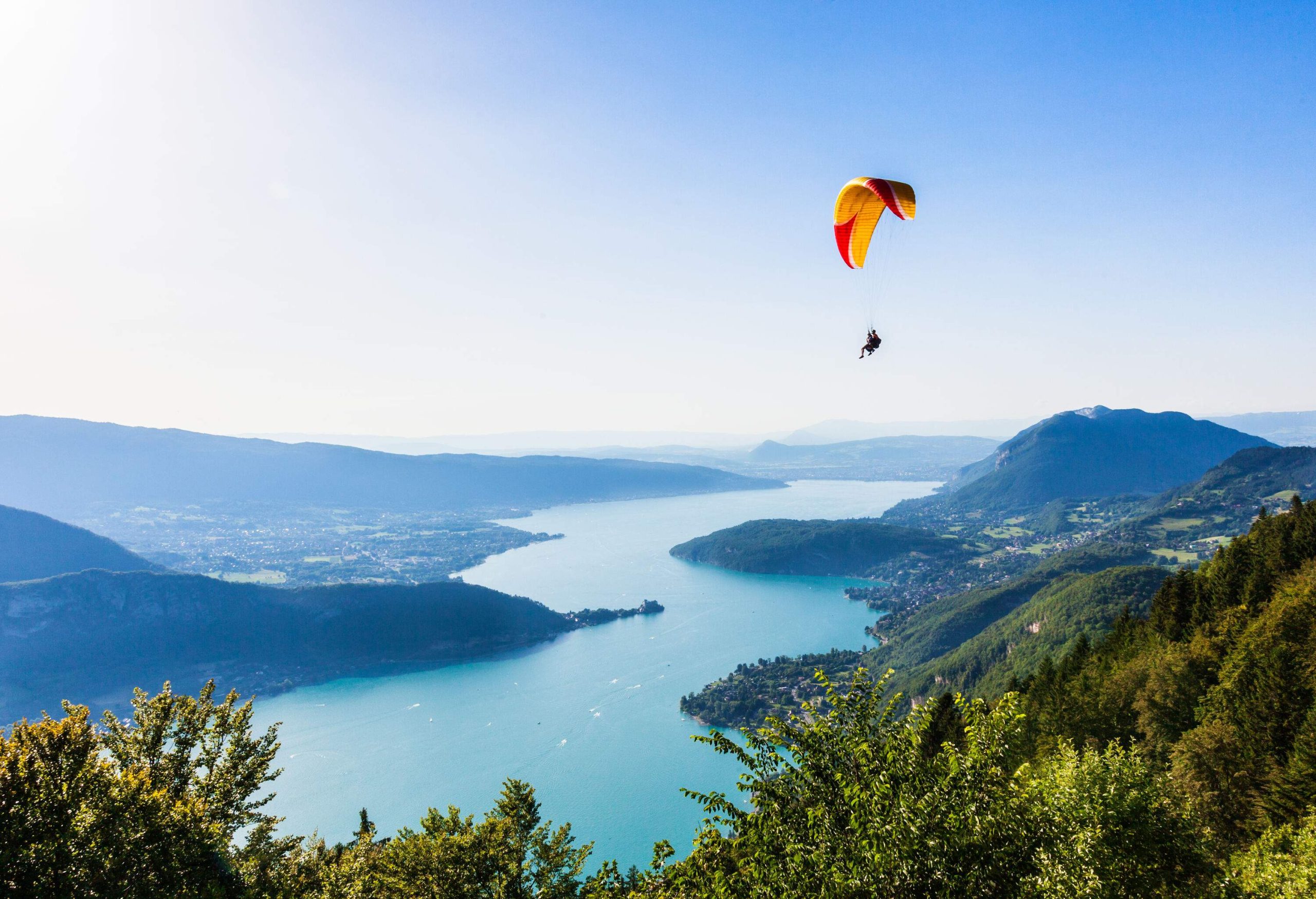 Two individuals are paragliding over a tranquil lake surrounded by forested islands.