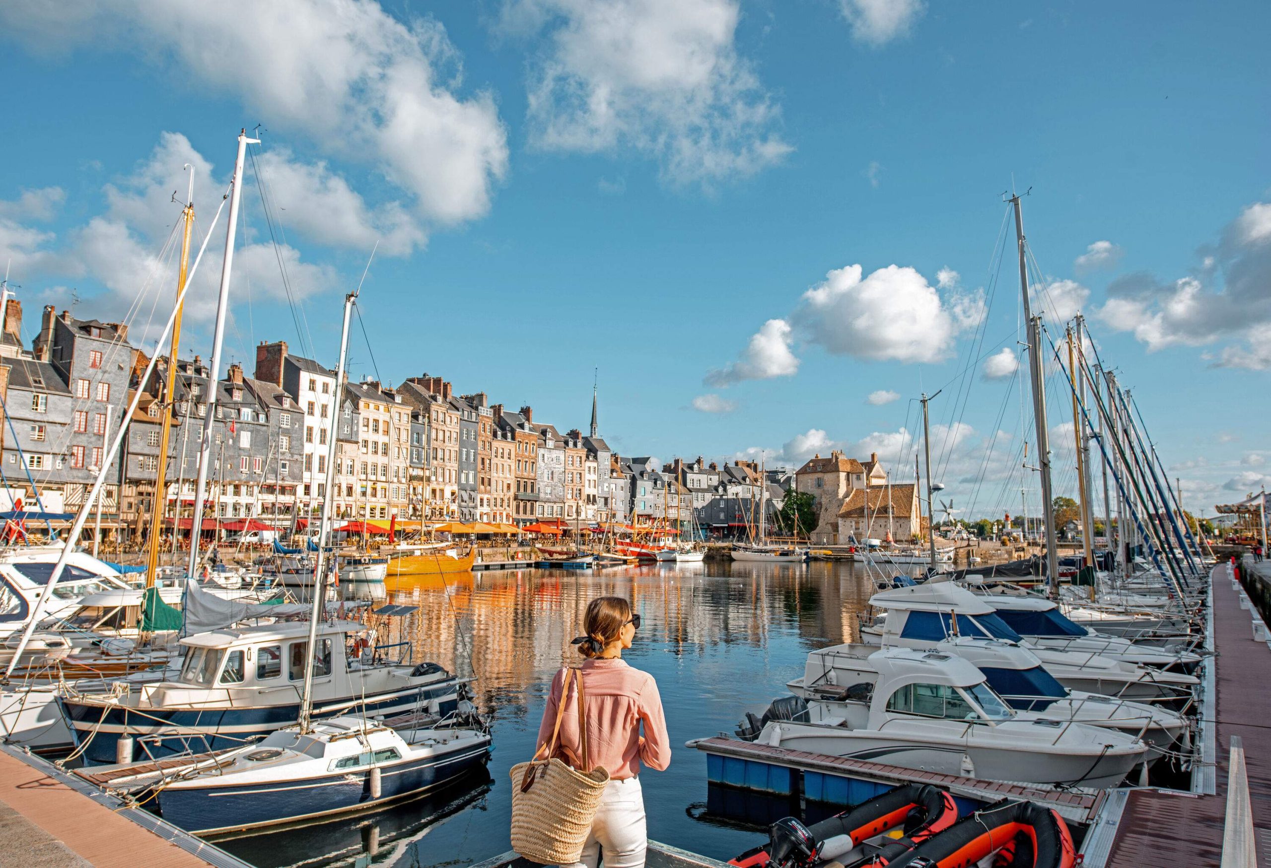 A woman with a rattan bag stands on a pier overlooking a marina full of docked boats.