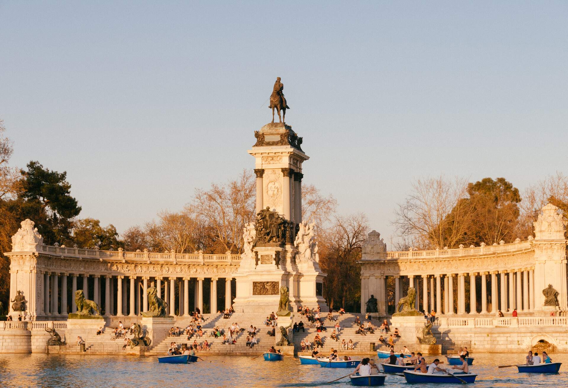 A monument of a horse rider on a large pedestal with other sculptures below it is backed by colonnades with steps leading to a pool where people paddle boats.