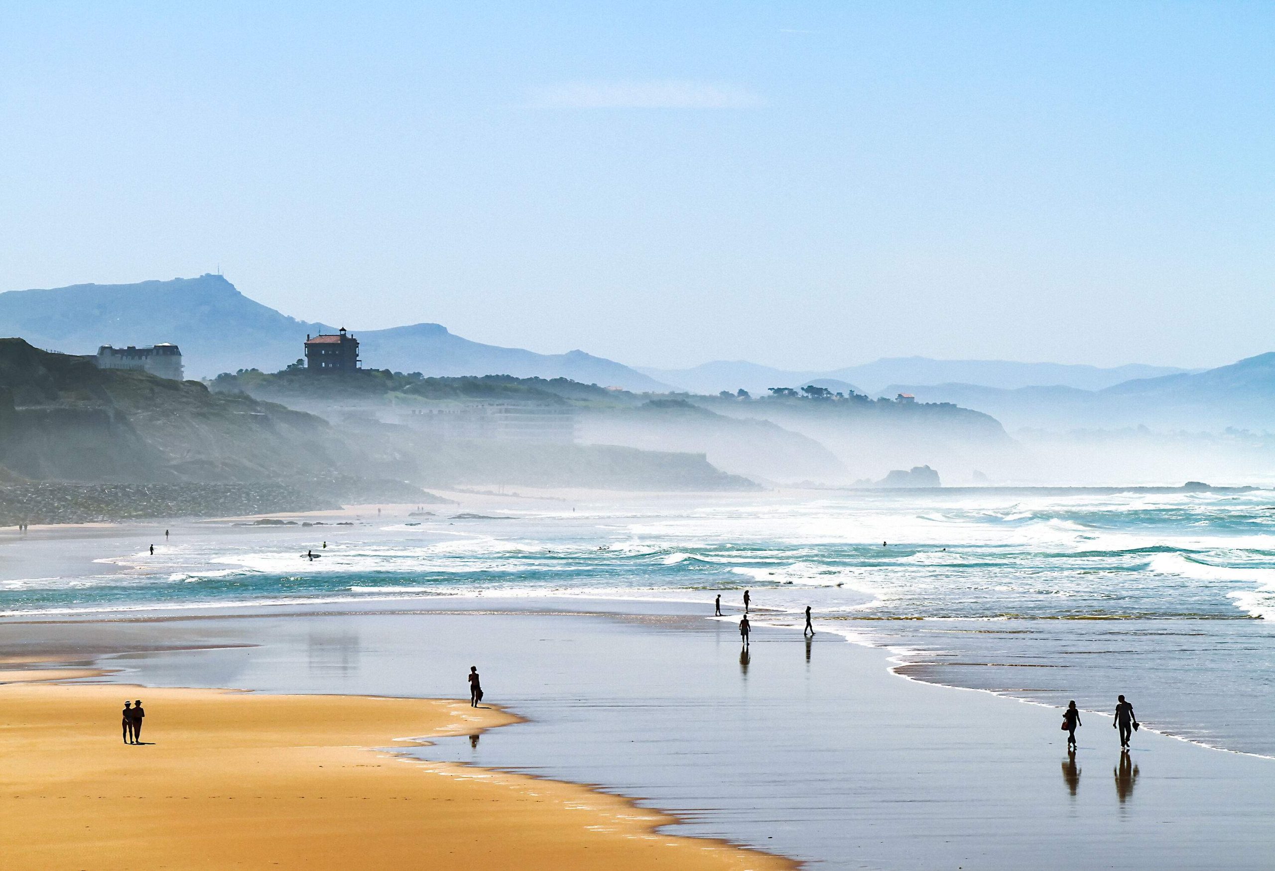 A silhouette of beachgoers against a background of cliffs and a mountain range shrouded by a hazy mist.