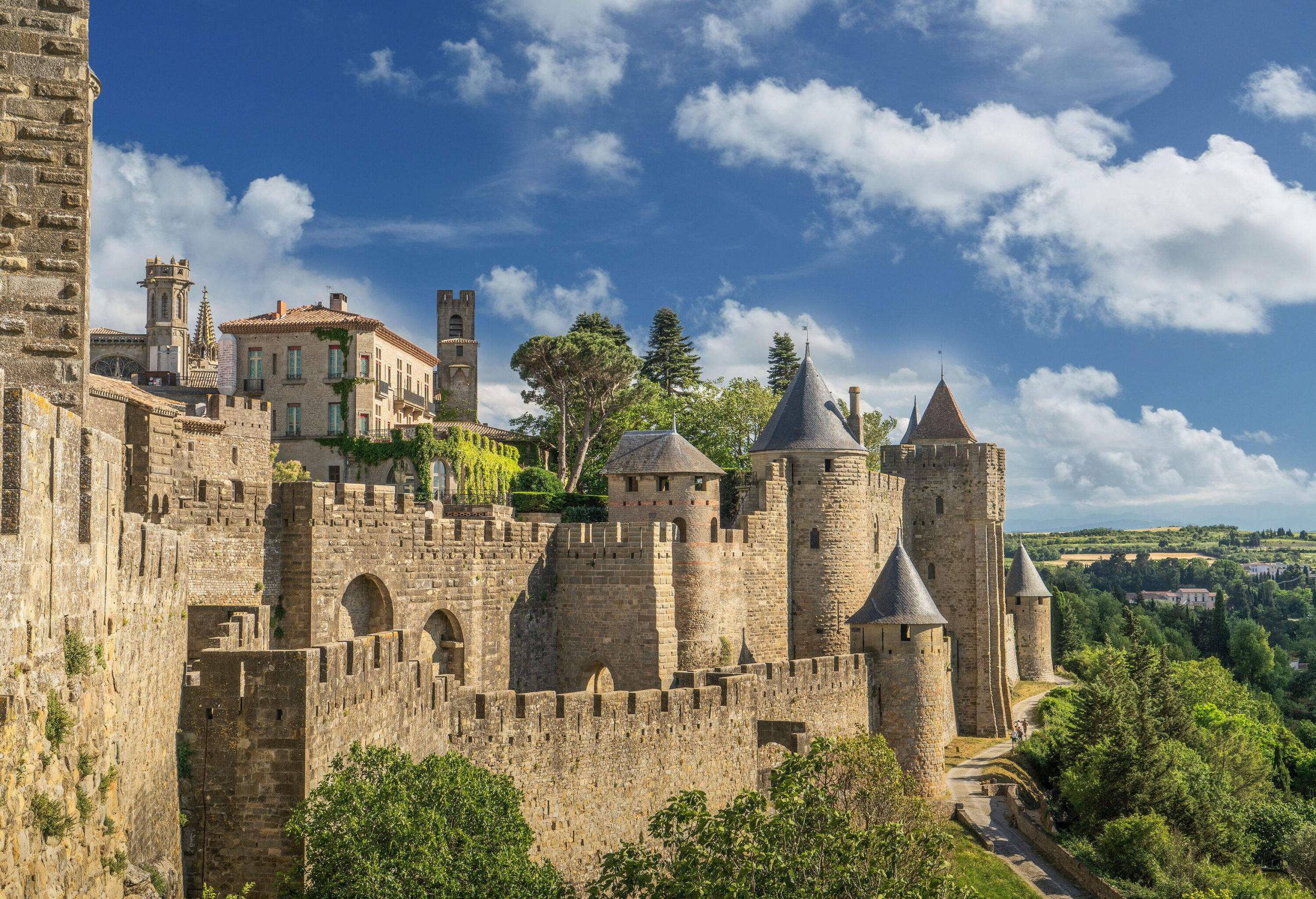 A multilayered wall with towers of a castle alongside a promenade on a lush green hill.