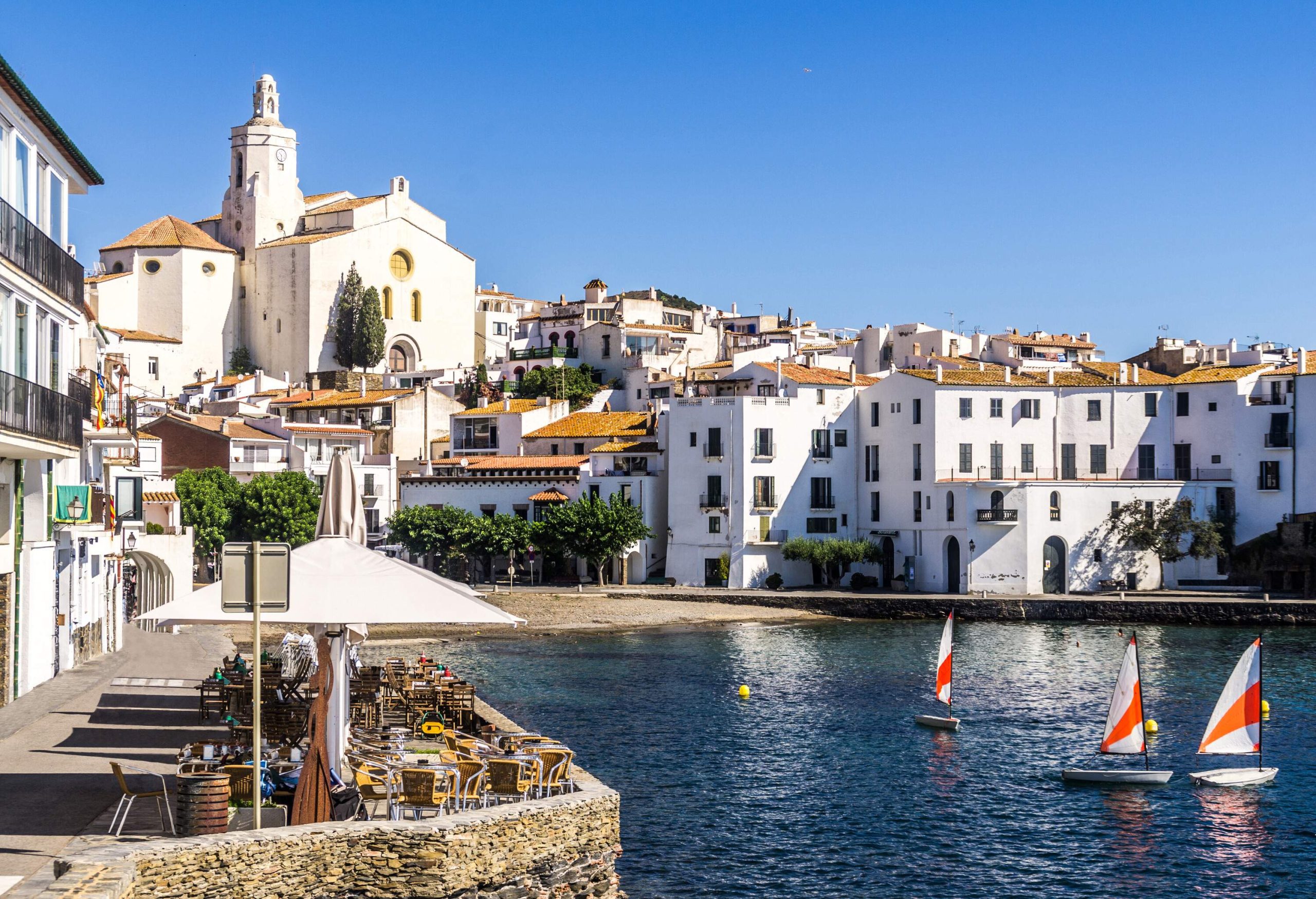 A church with a bell tower overlooks the white buildings of a town along a coast with outdoor seating areas.