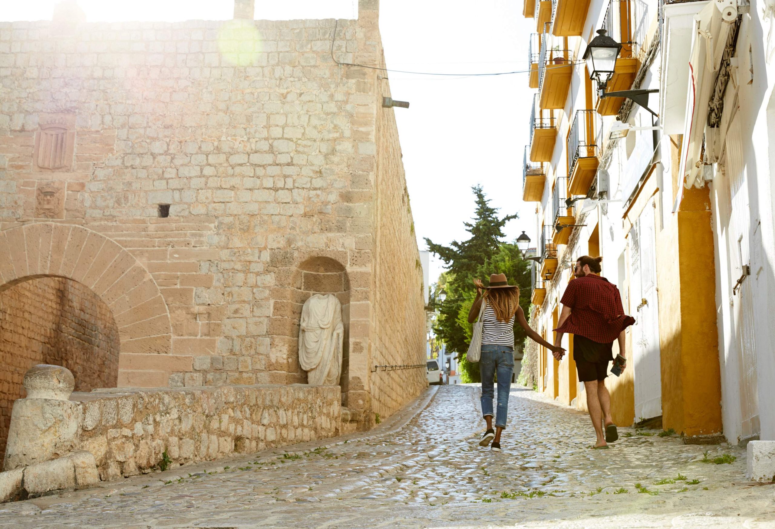 A couple in casual clothes walks on a cobblestone street between the neighbourhood buildings.