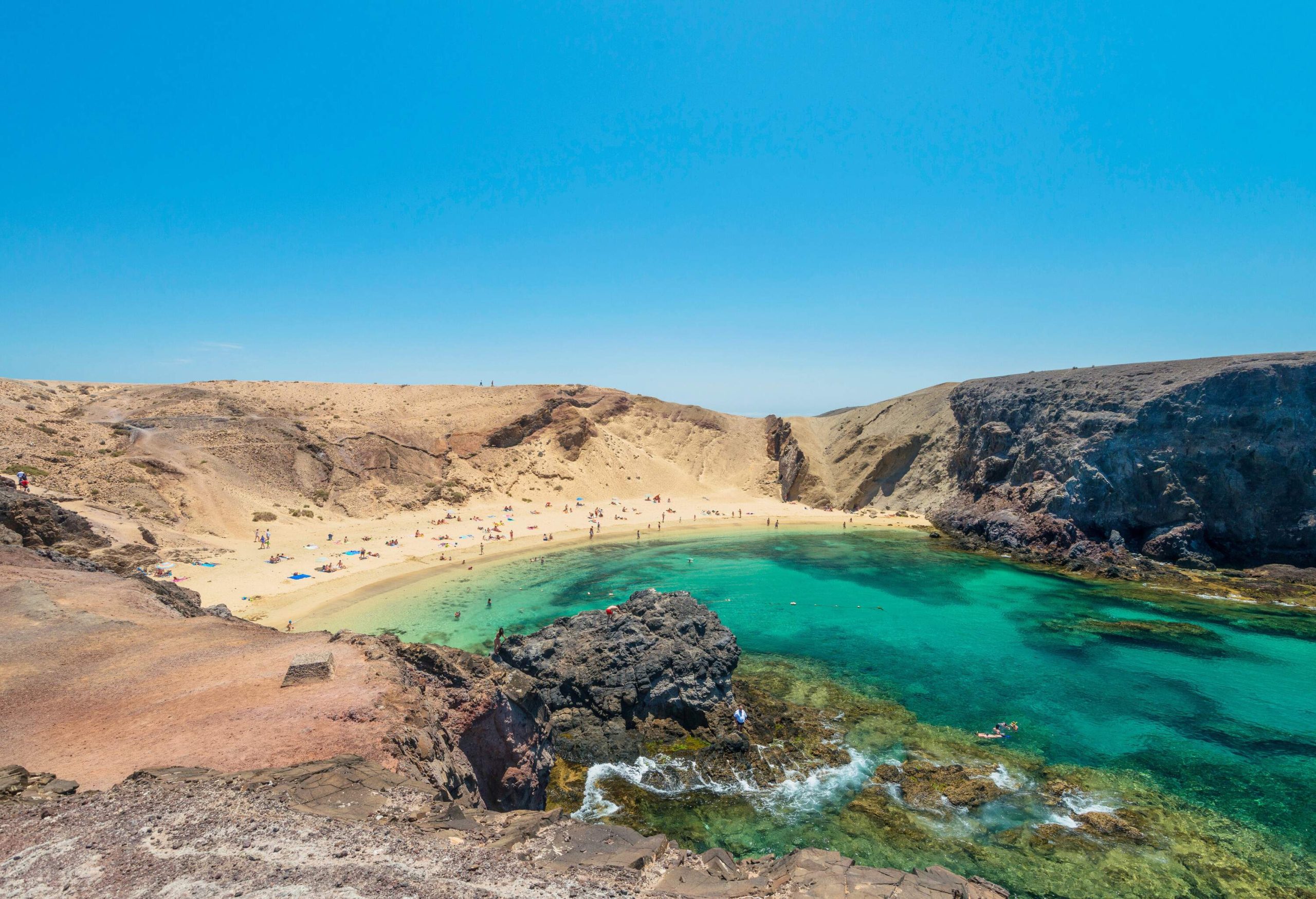 People on a cove of white sand beach with shallow clear emerald green water bordered by rocks.