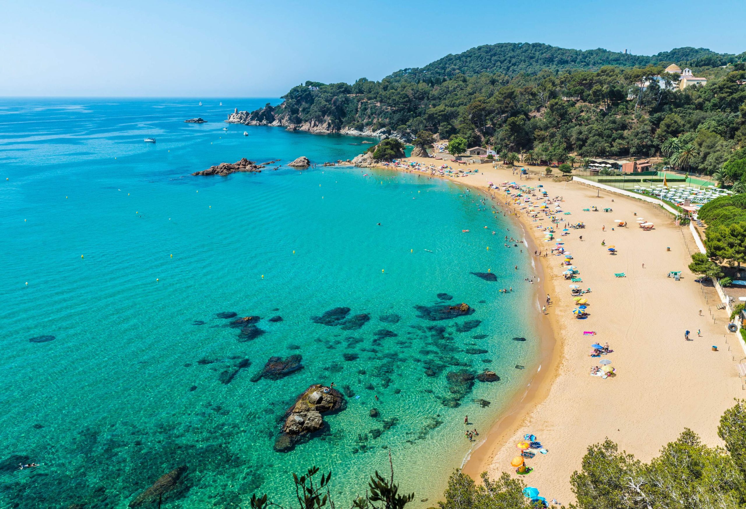 A hilly coastline with forested slopes and seashore establishments surrounds a beach with jutting rocks.