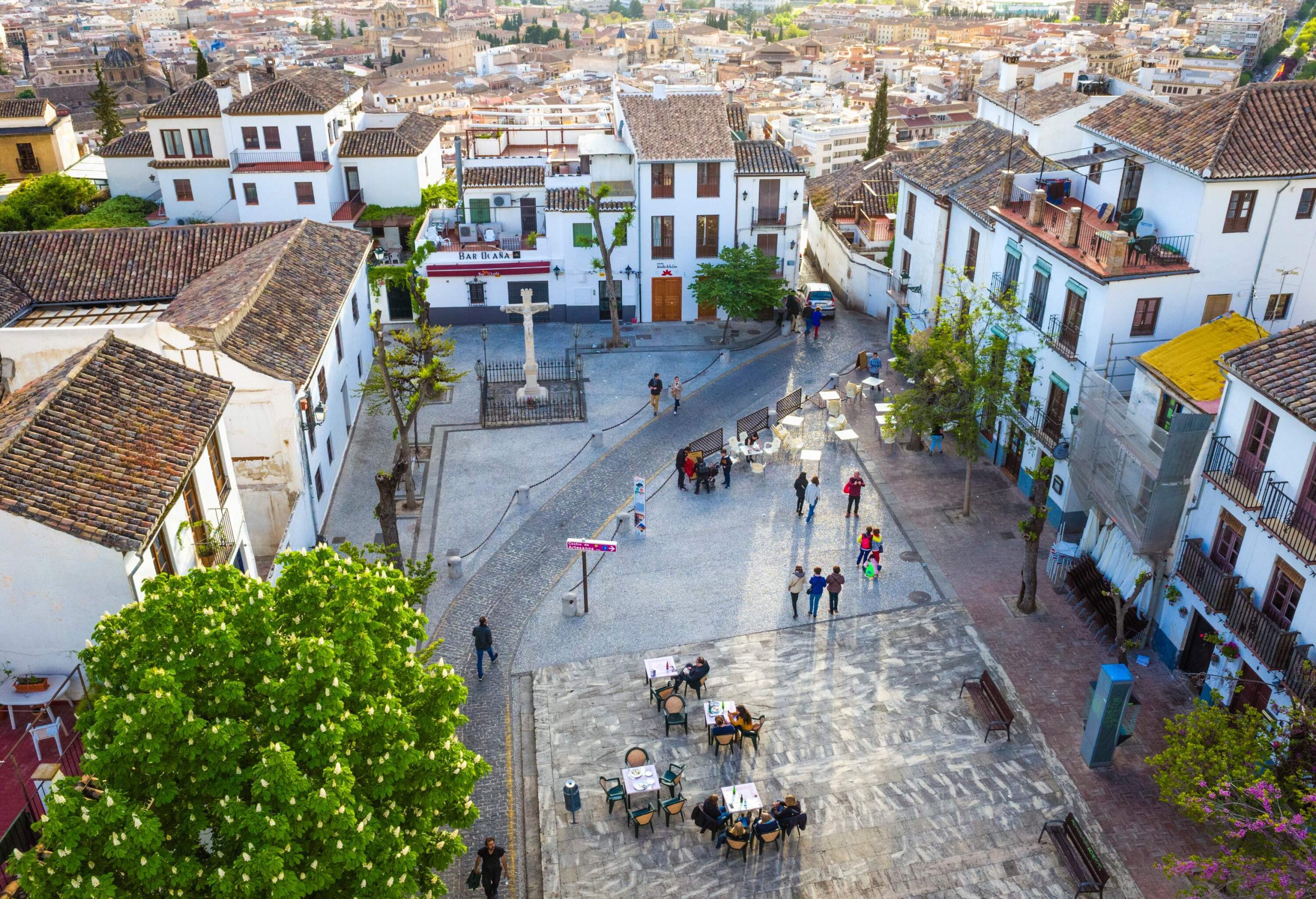 A simple public square with individuals sitting at outdoor cafes and others walking between row houses.