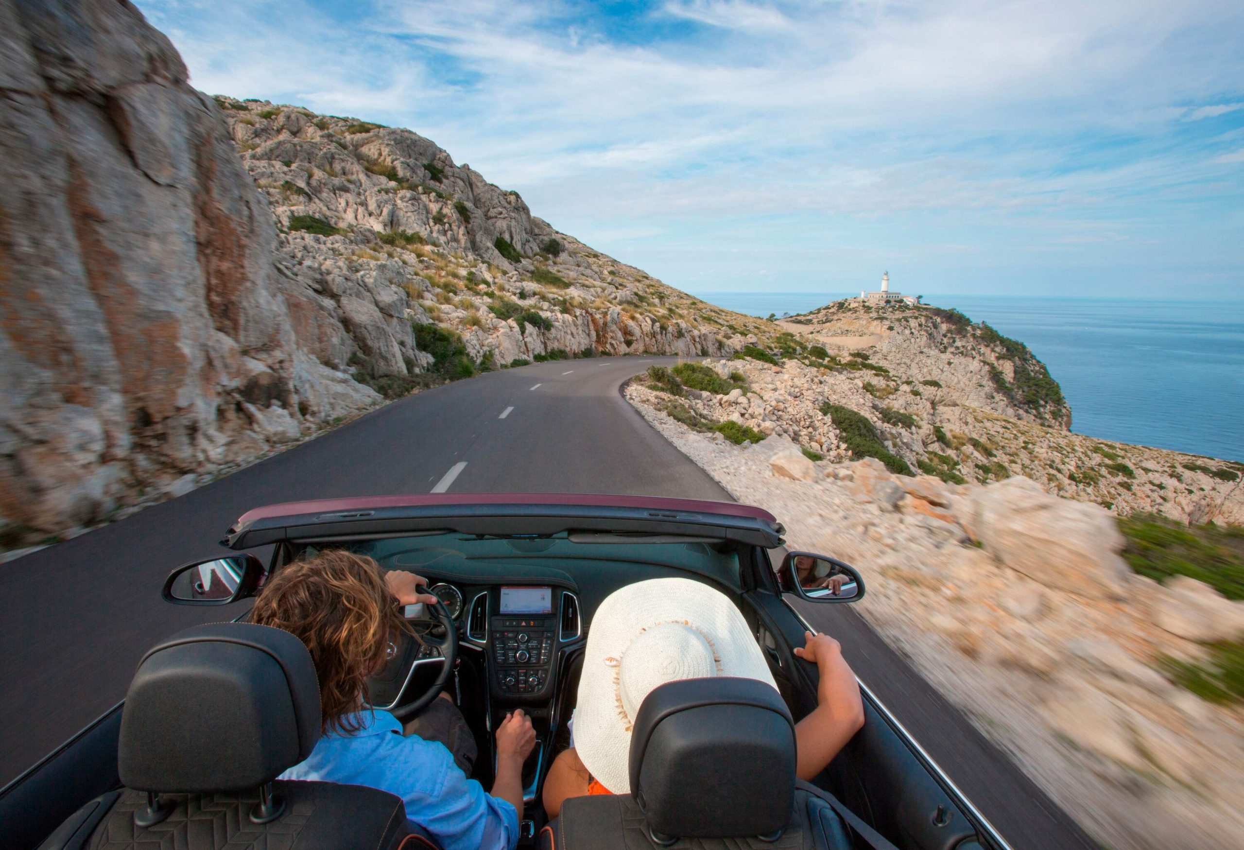 Two people in a convertible on a rocky mountain road overlooking a white lighthouse on the edge of a cliff by the sea.