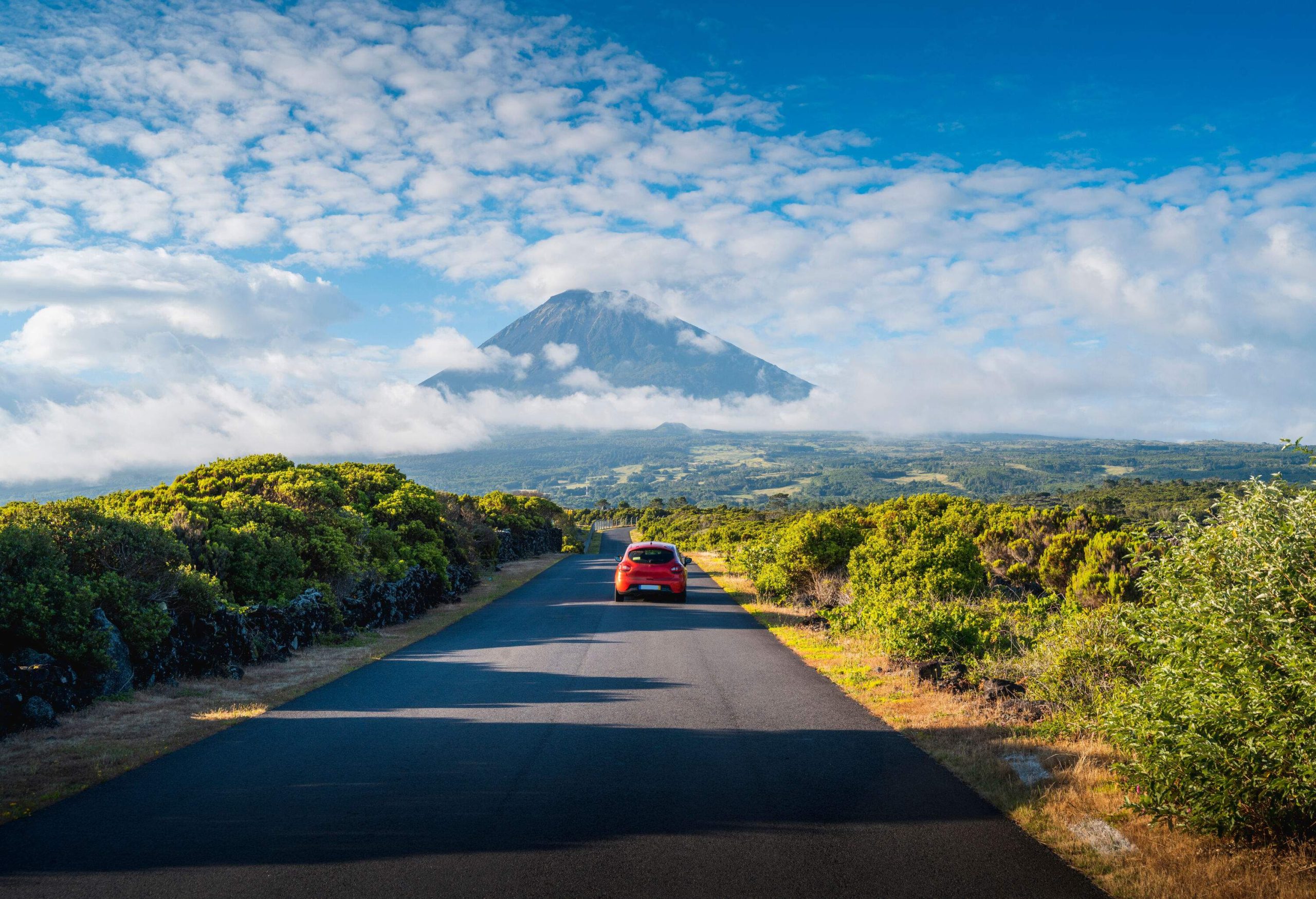 A red car travels on a road across a verdant landscape with distant views of a mountain covered in clouds.