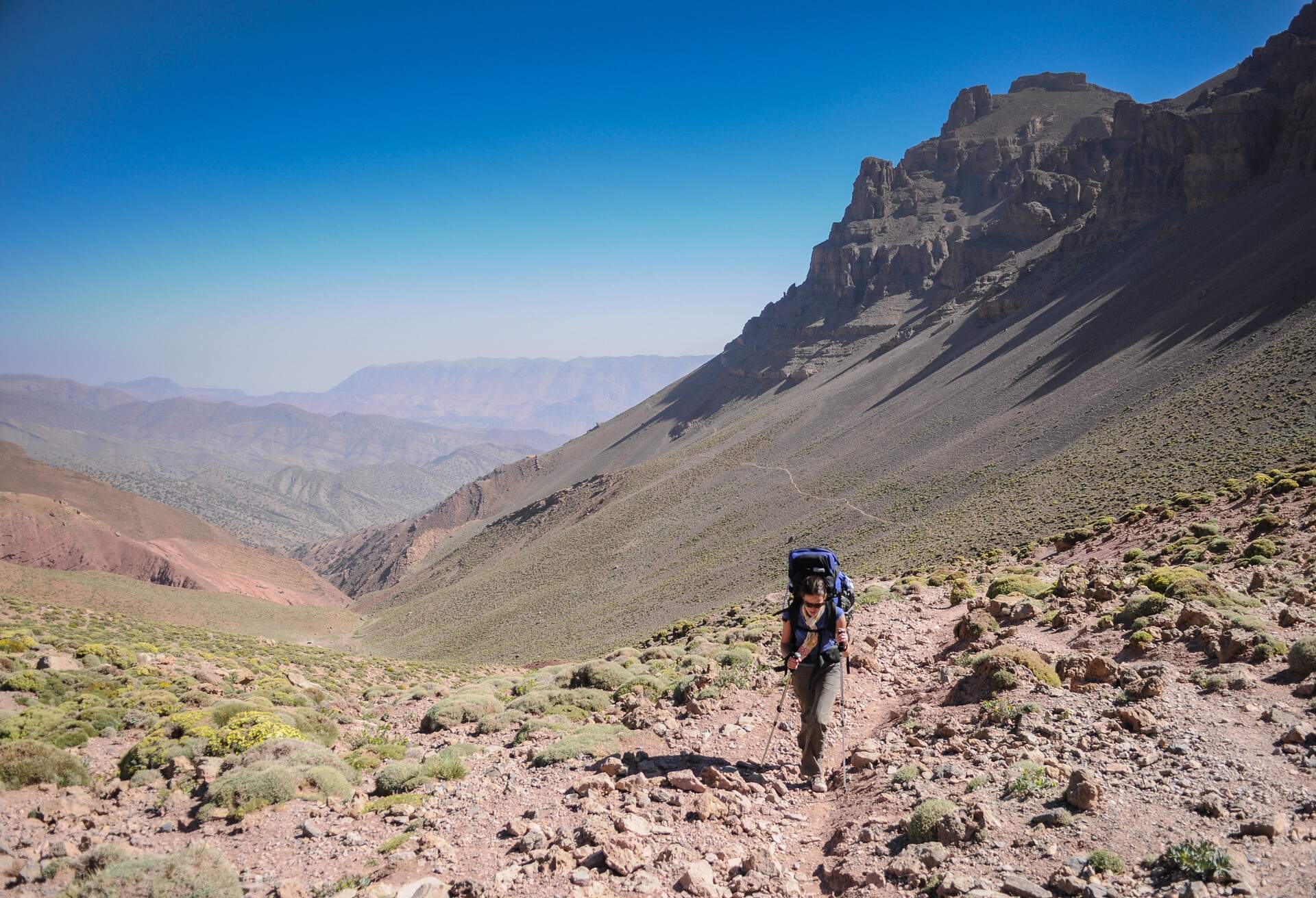 Female Hiker walking up a mountain pass with backpack on her back on a single footpath up a mountain with a valley and a clear blue sky behind Atlas Mountain Toubkal Morocco