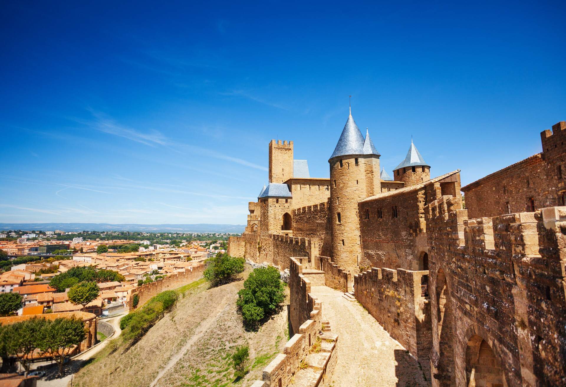 The walls of a castle with a pointed roof tower atop sloping ground and overlook the settlement below.