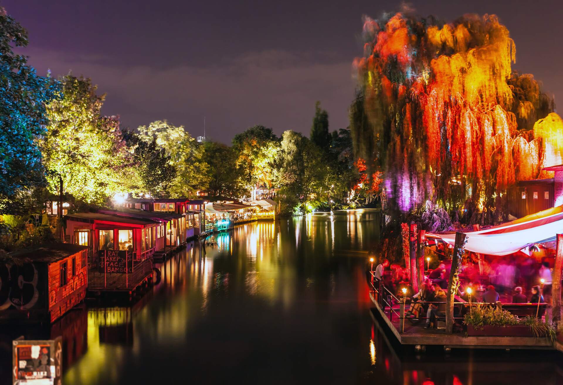 A night-time view of a calm river bordered by commercial establishments, colourful illuminated trees, and a crowded riverside bar with a terrace.