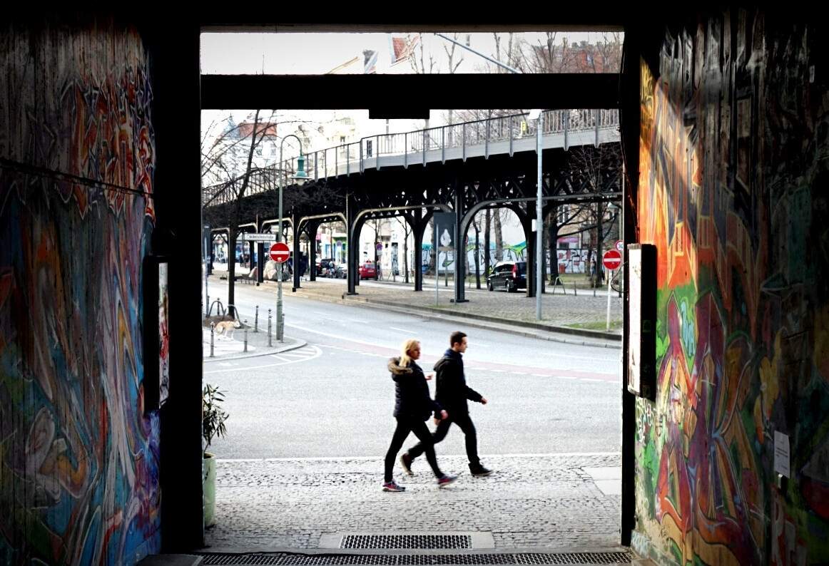 Two people walk past the entrance of a street with walls painted with colourful graffiti.