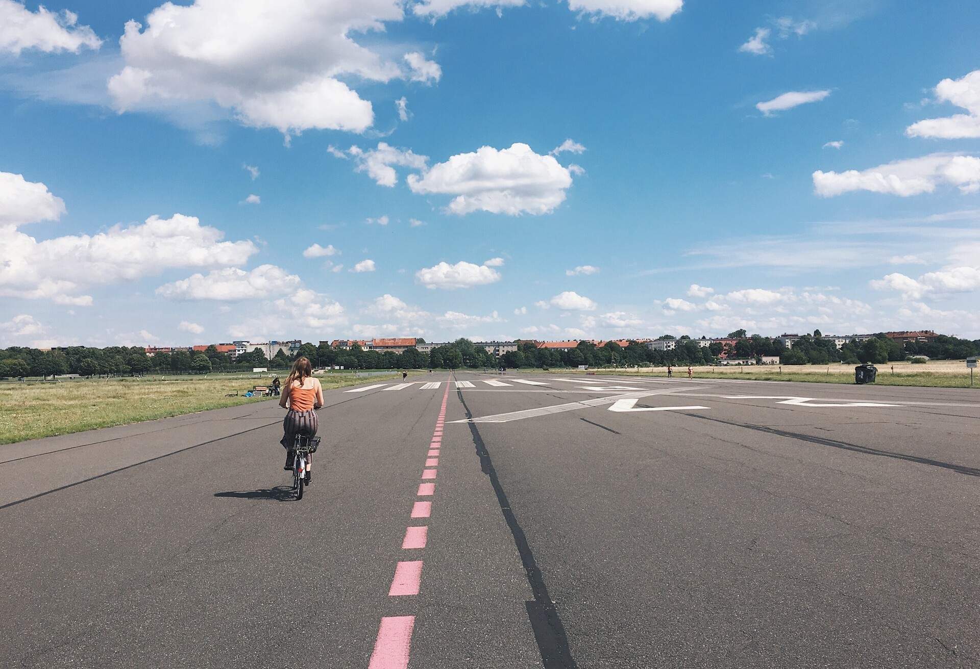 A woman cycling along a cobblestone road, admiring lush trees and a cloudy blue sky.