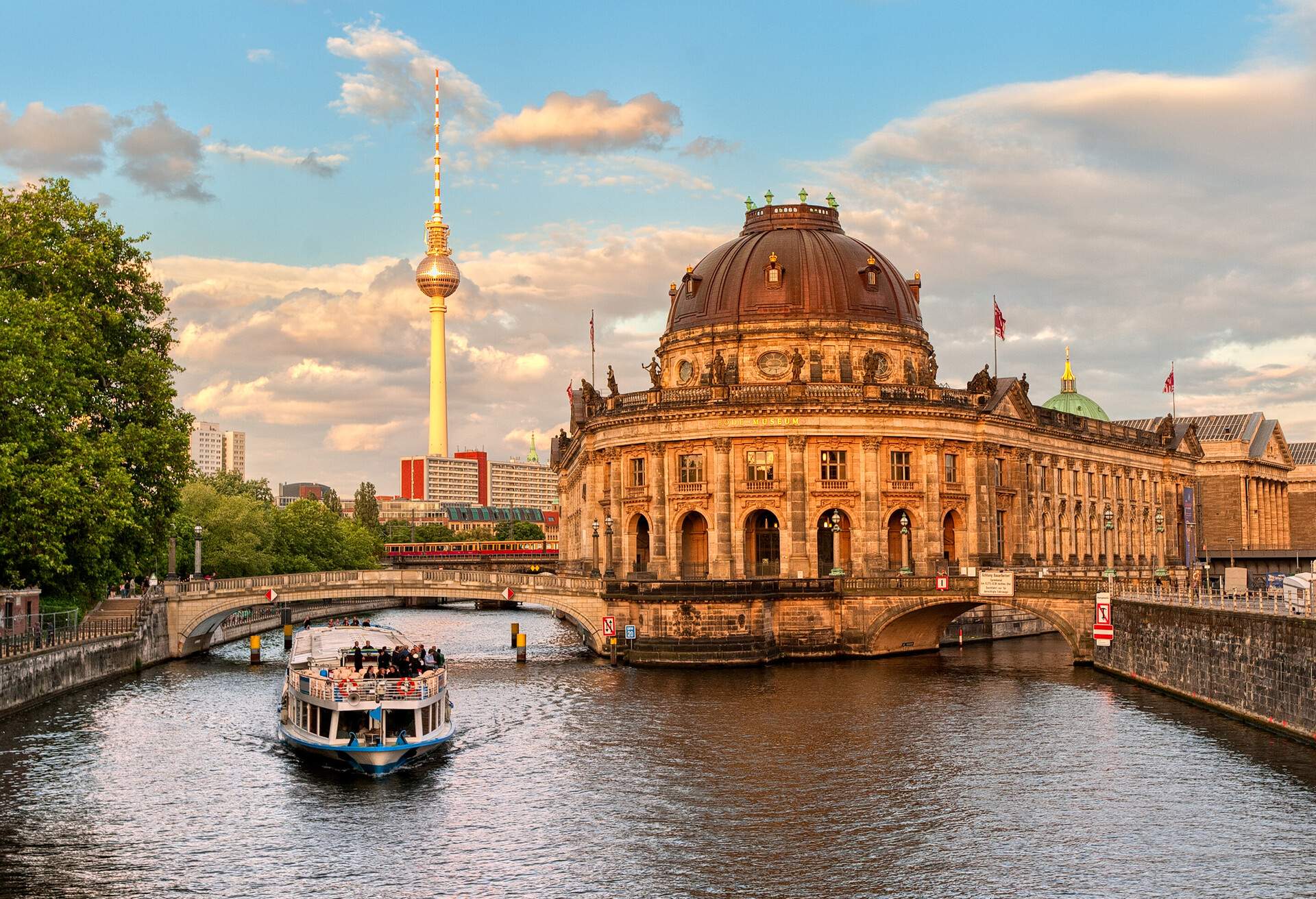 A passenger boat cruises a river with views of the Baroque-style Bode Museum and the Berlin TV Tower.