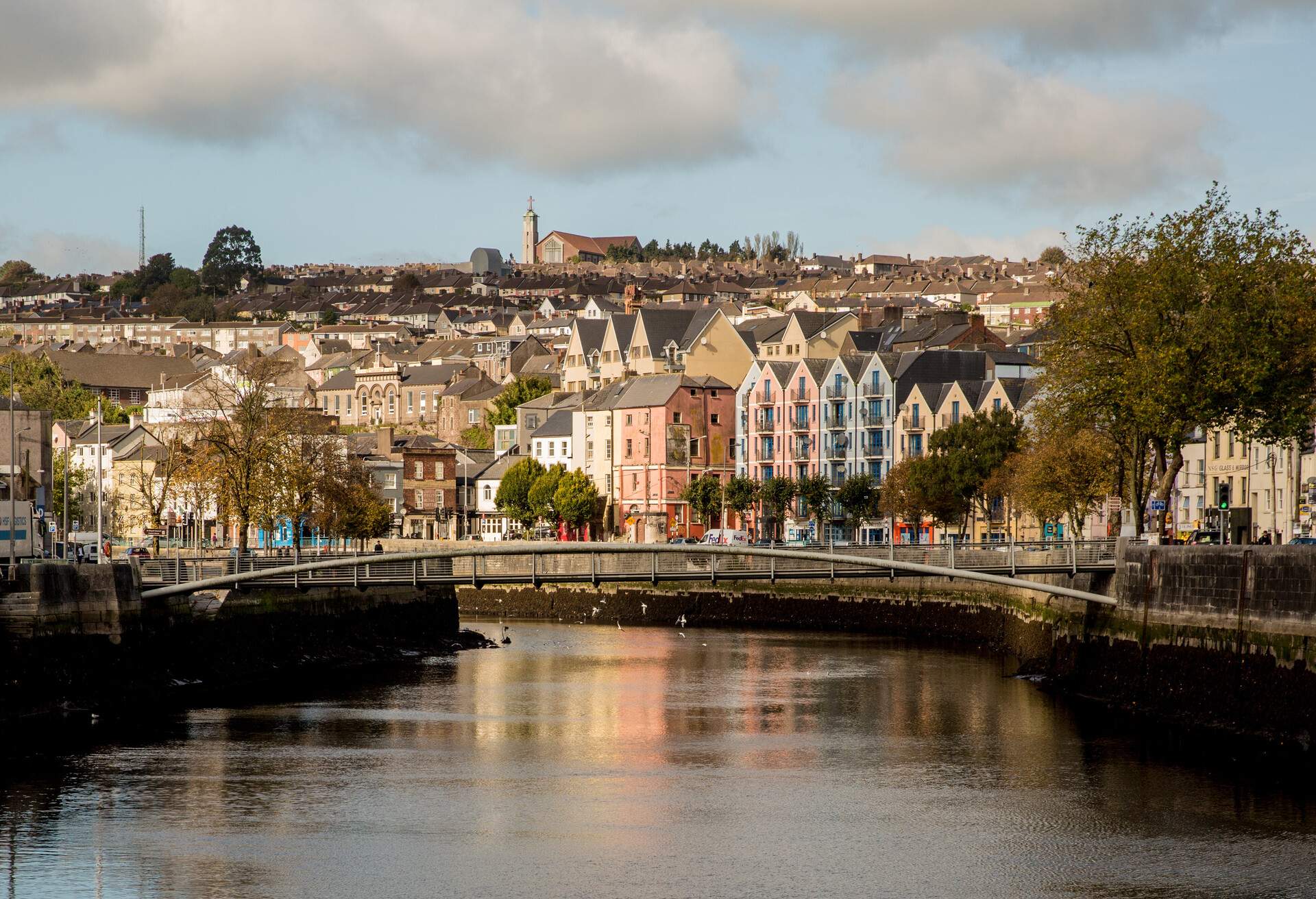 A broad river passes through a narrow footbridge alongside a thick cluster of compact tall buildings layered on a slope.