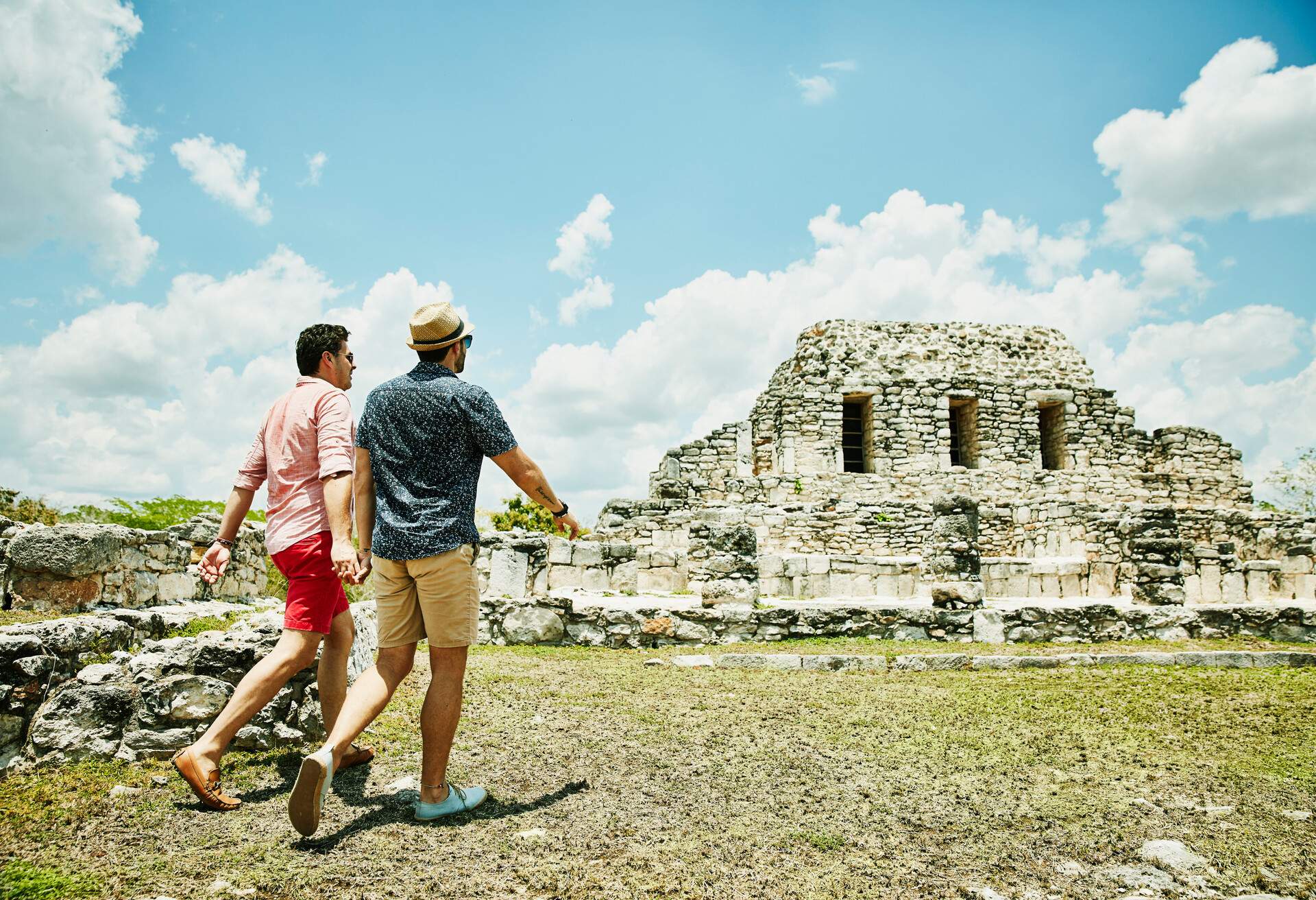 Two men holding hands while walking towards a historical Maya ruins.