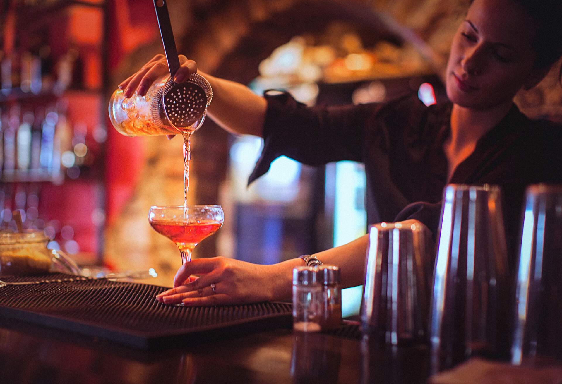Close-up of a young female bartender pouring cocktail in a nightlife cocktail bar. Selective focus. Focus on foreground.