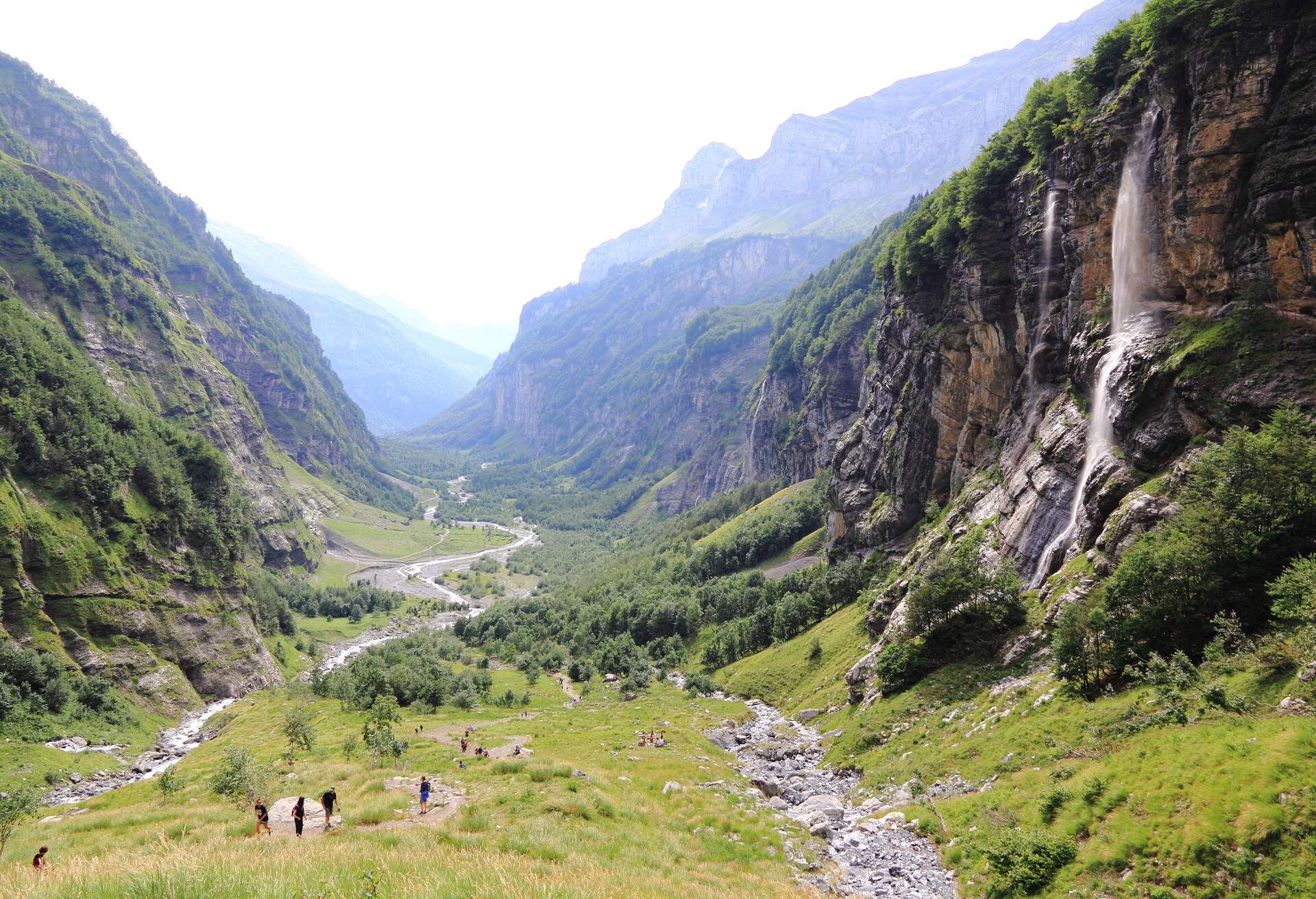 A valley with a river and huge mountains on both sides, one flank features a waterfall.