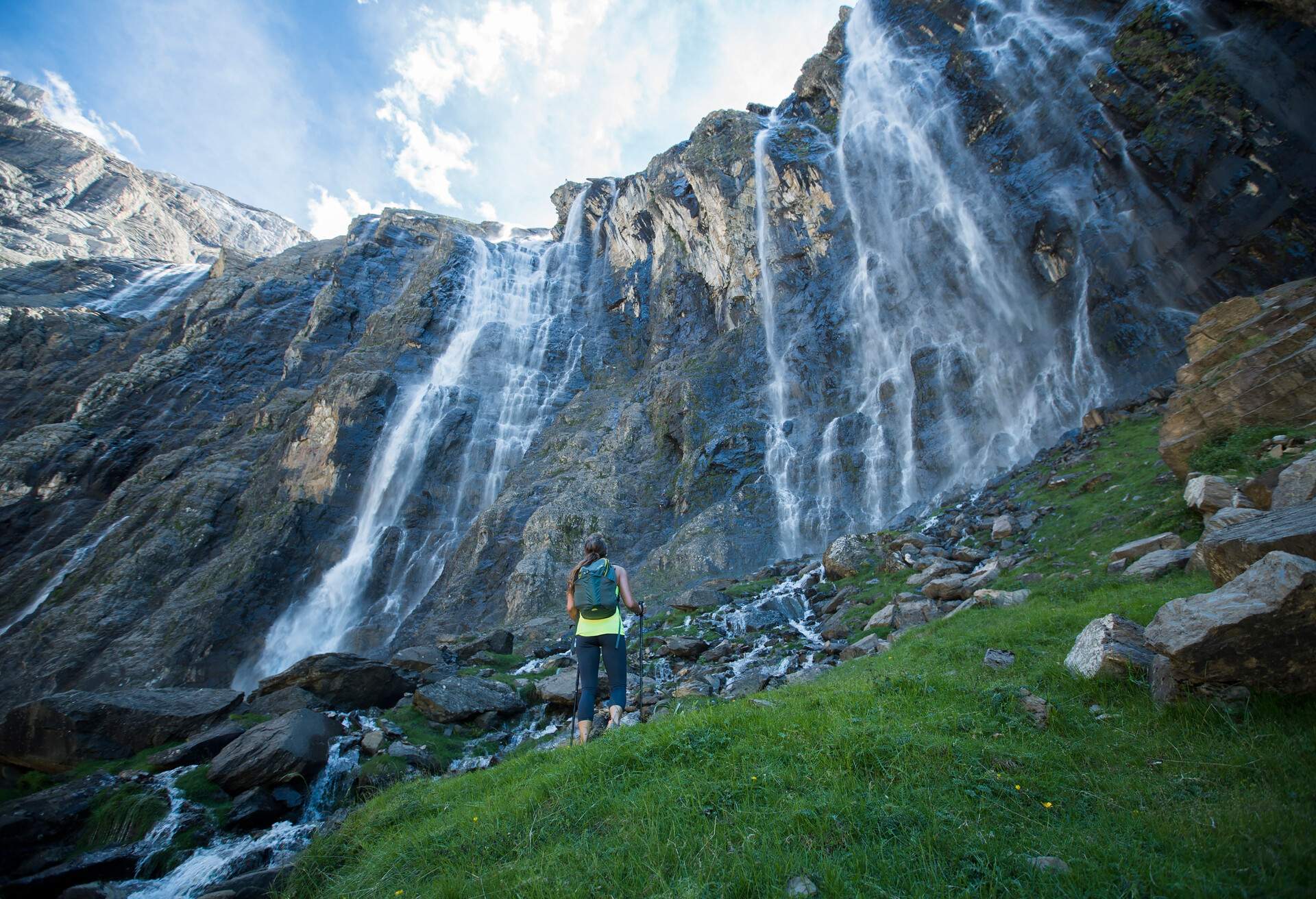 A woman holding hiking poles gazing up at the spectacular rush of a waterfall.