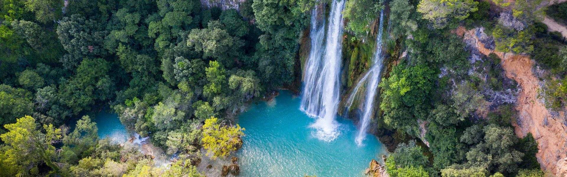 A thick forest surrounds a waterfall that flows into a calm river.