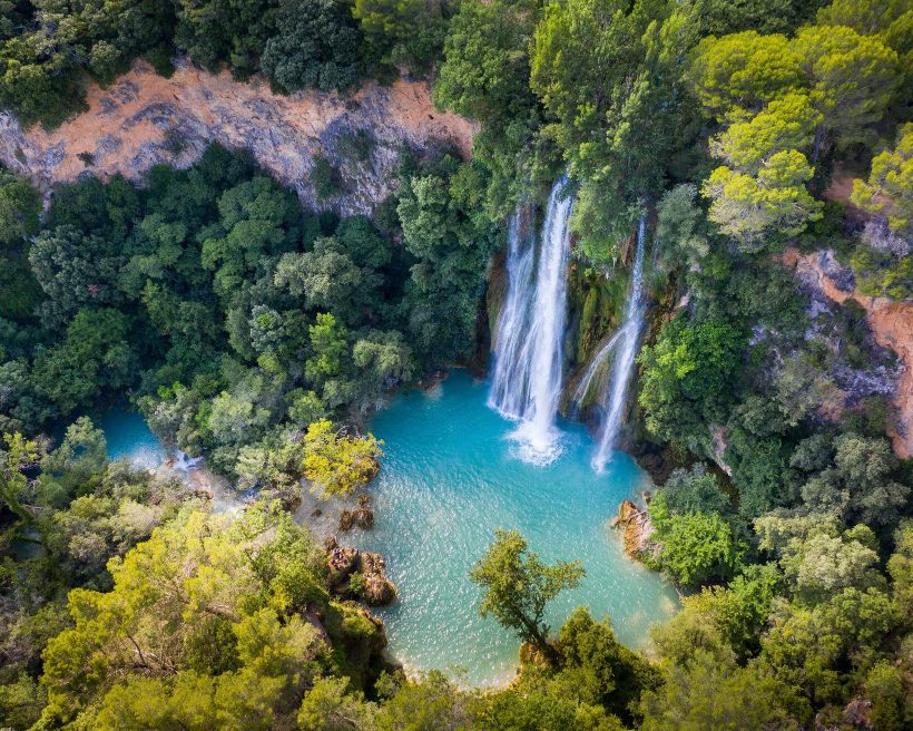 A thick forest surrounds a waterfall that flows into a calm river.