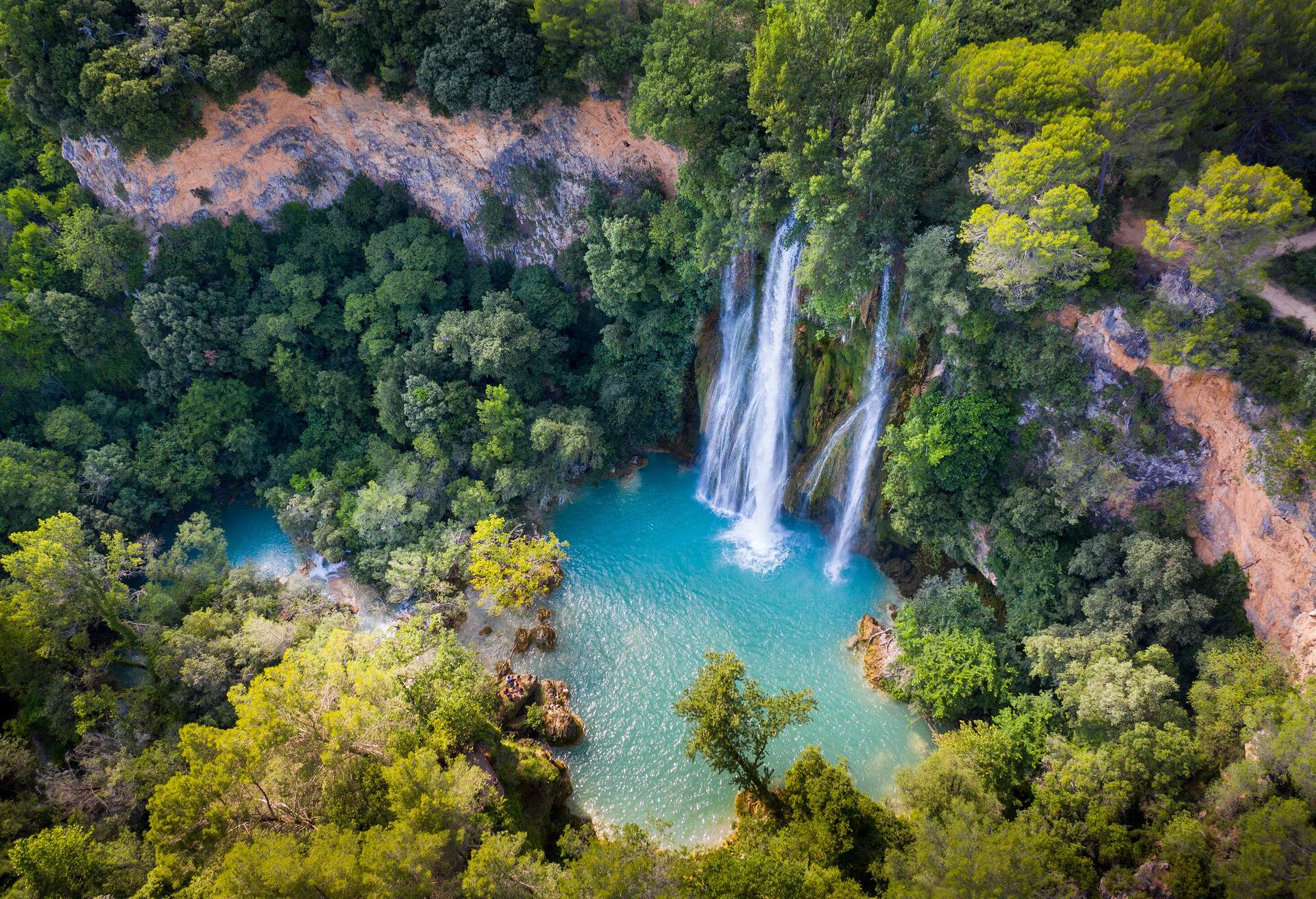 A thick forest surrounds a waterfall that flows into a calm river.