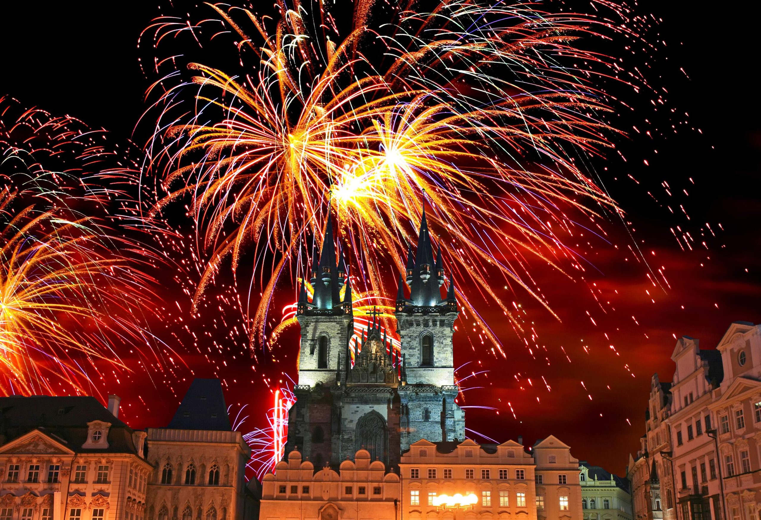 A night-time view of a crowded public square surrounded by illuminated city buildings and a church with pointed twin bell towers under the colourful exploding fireworks.