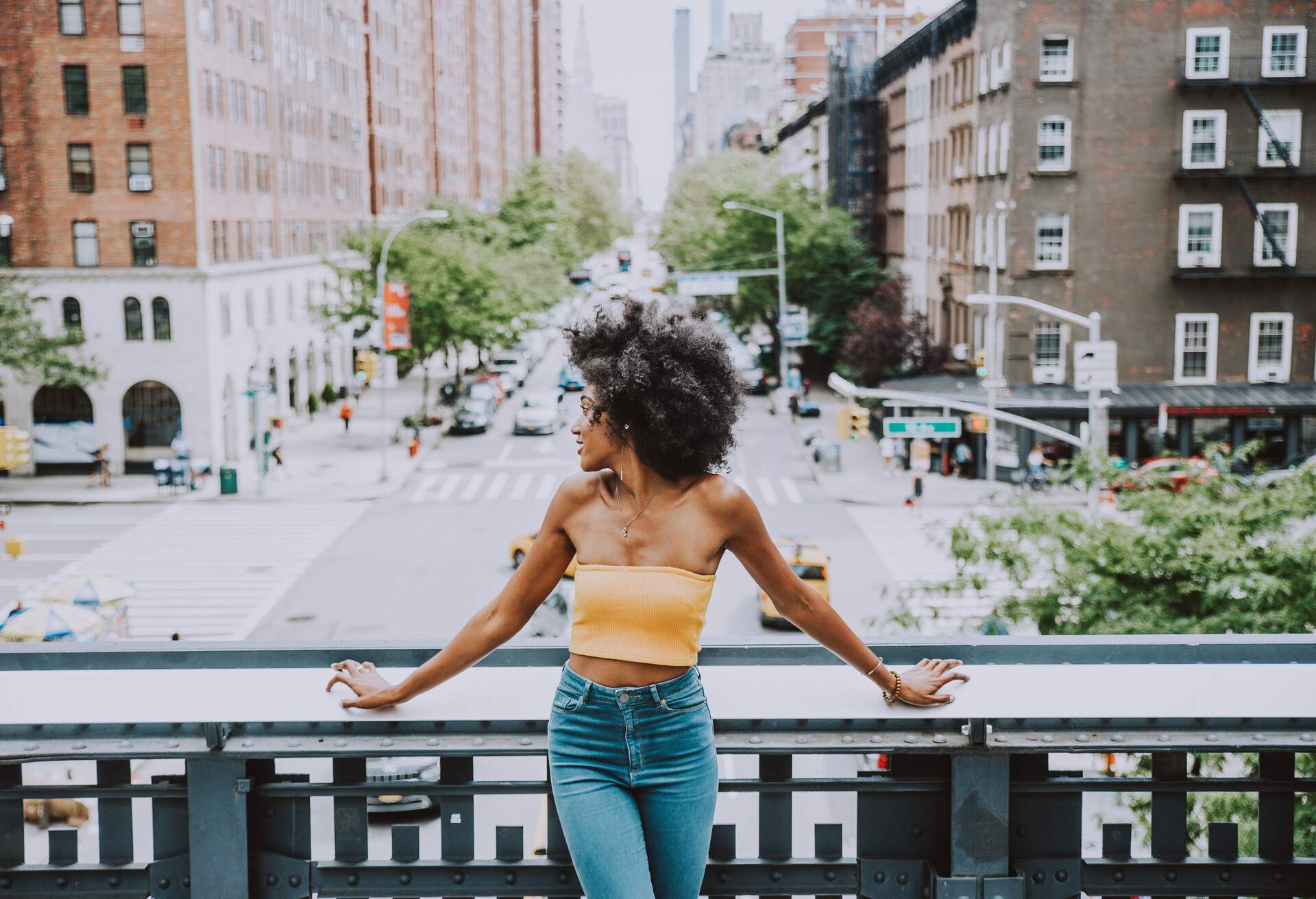 A curly-haired woman in a yellow tube top and denim jeans leans on a bridge railing as she looks sideways.