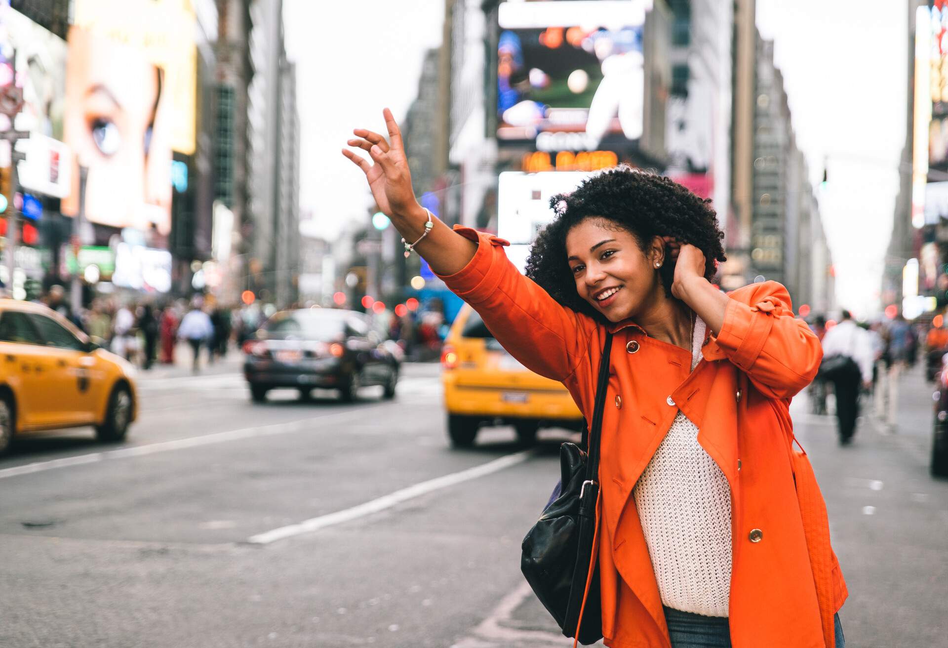 A curly-haired woman in a striking orange coat waves her hands for a cab along a busy street.