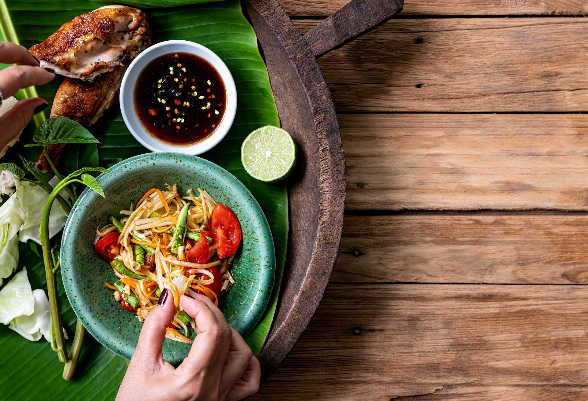 Thai lady eating traditionally with her hands, fresh world famous Som Tam (papaya salad) with BBQ chicken, sticky rice and raw salad vegetables on an old wooden table background.  Good copy space to the right of the image.