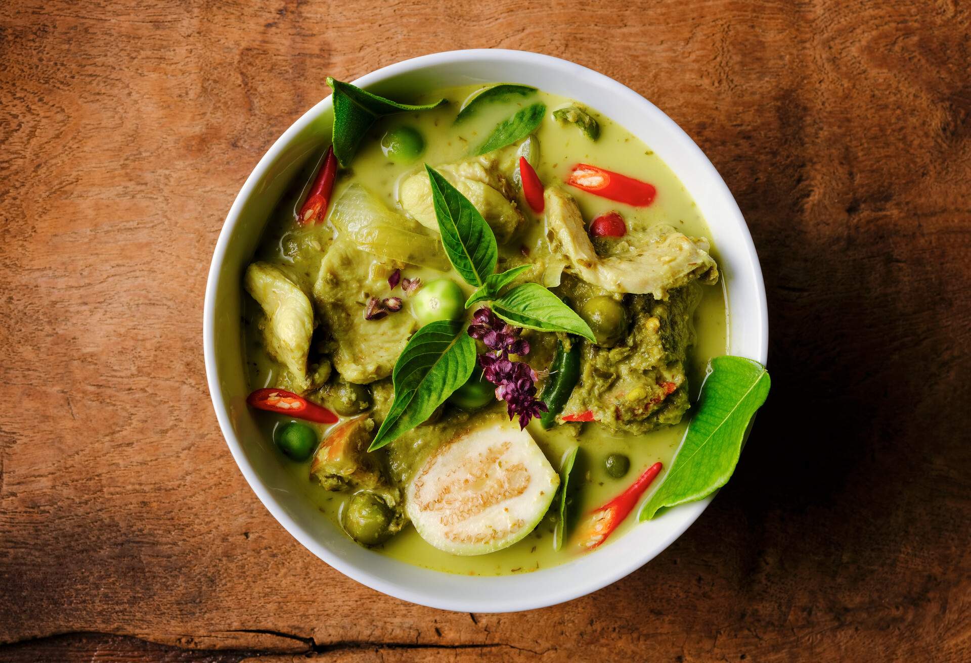 This Thai known in Thai as 'Gaeng Keow Wan Gai' dish is very famous and known all over the world and one of Thailand's popular signature dishes when it comes to Thai food. This dish consists of ingredients including a green herb-based base with fresh coconut milk, sweet basil, Thai aubergines, pea aubergines, chicken, and spicy fresh red chili. The combination of ingredients results in a sweet and spicy taste with a variety of complementary textures and is normally eaten together with fresh steamed Jasmine Thai rice or fresh rice noodles. The image was taken from directly above with the bowl set on an old worn textured wooden background with lots of grain and character.