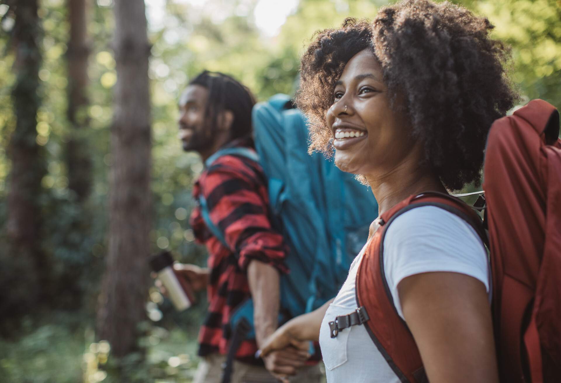 Smiling couple in a woodland holds hands and carries large hiking bags.