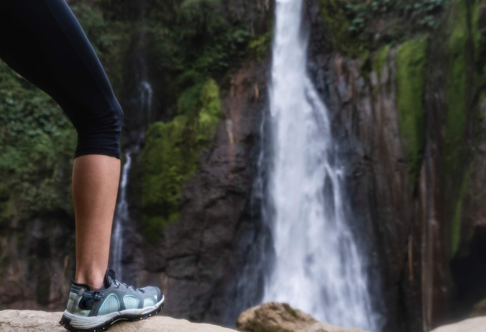 Selective focus on the leg of a woman standing in front of a wild waterfall