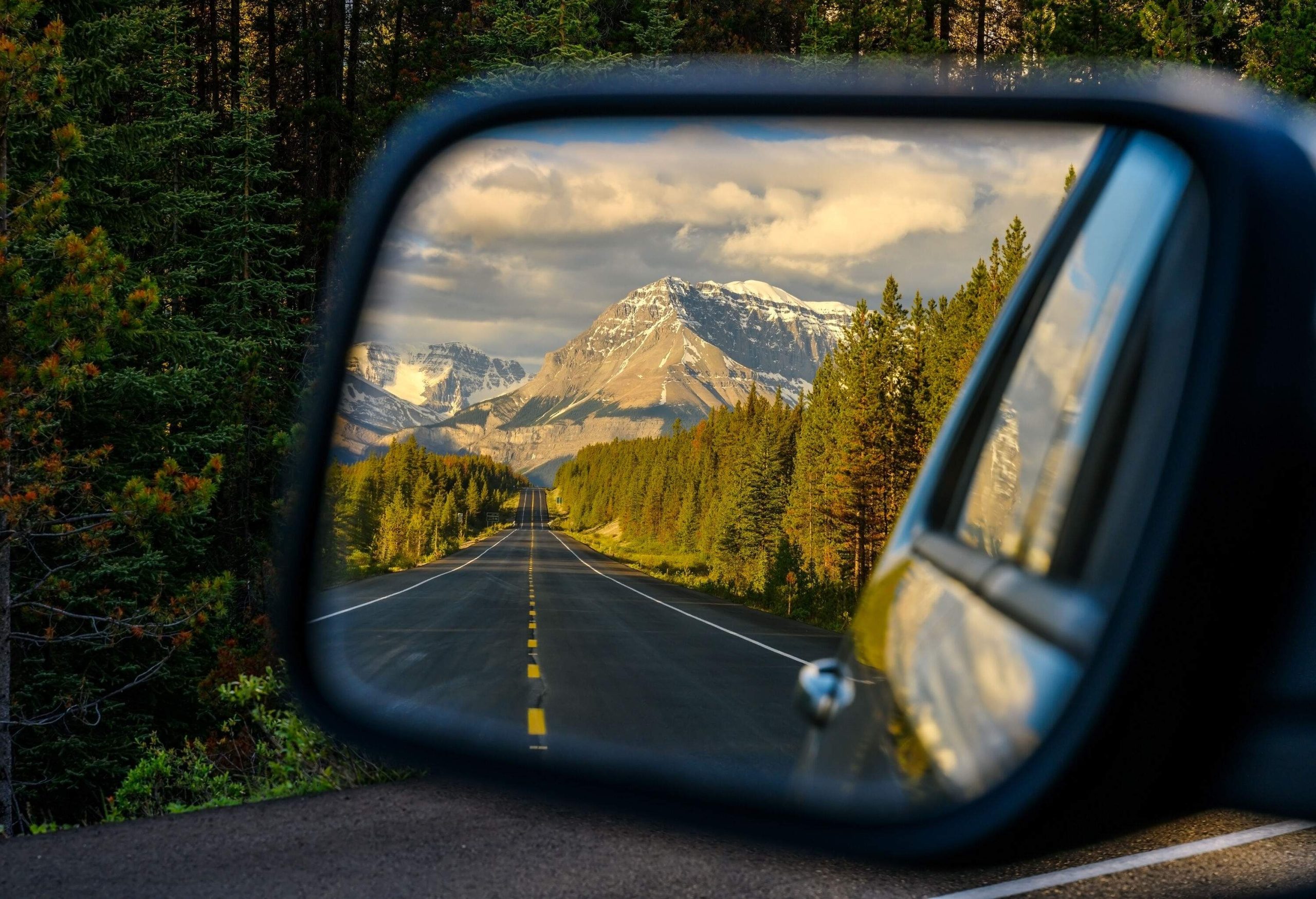 A car's side mirror with a reflection of an empty highway across a forested landscape with a snow-capped mountain in the background.